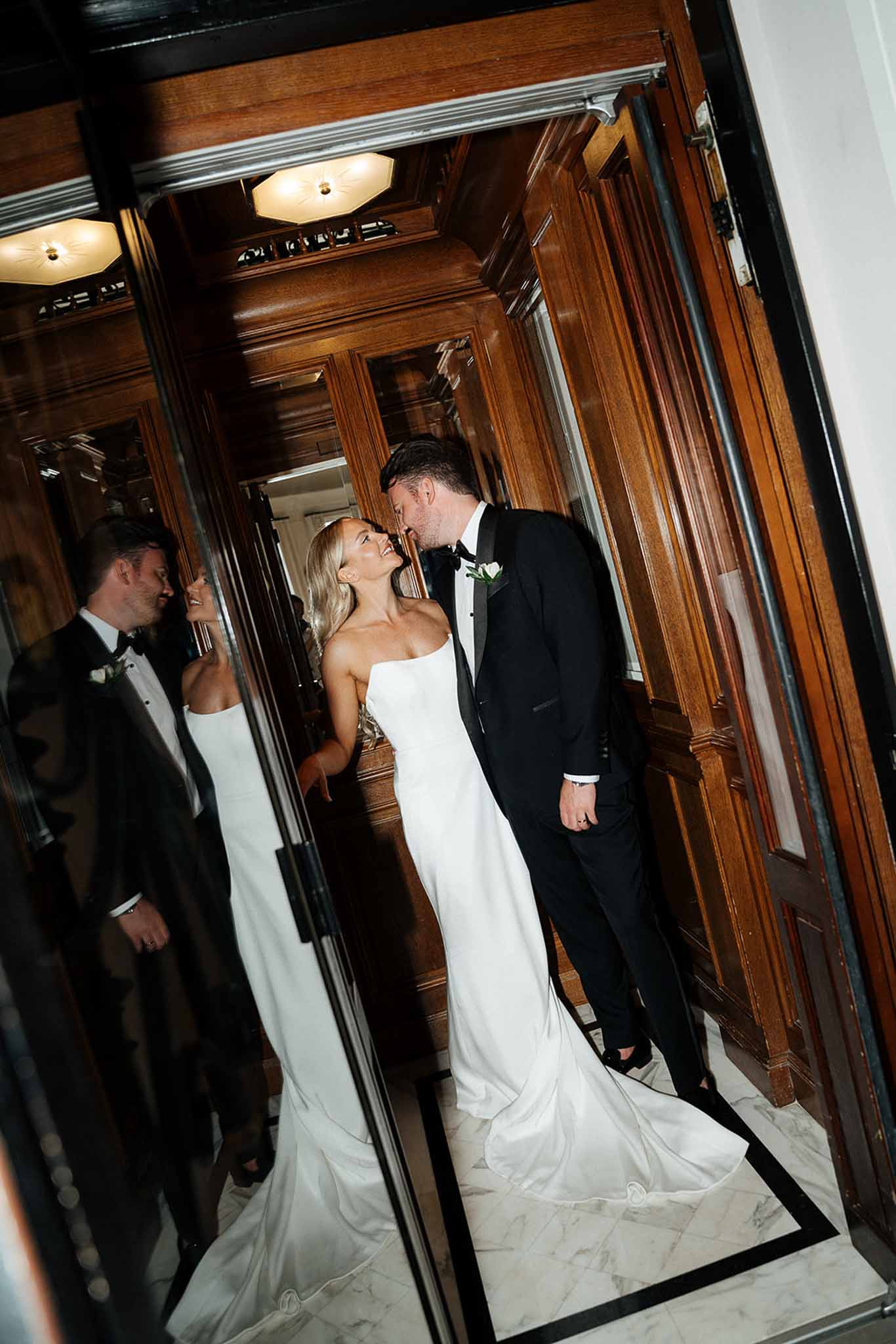 Bride and groom leaning in to kiss inside wood-paneled hotel elevator with mirrored reflection visible
