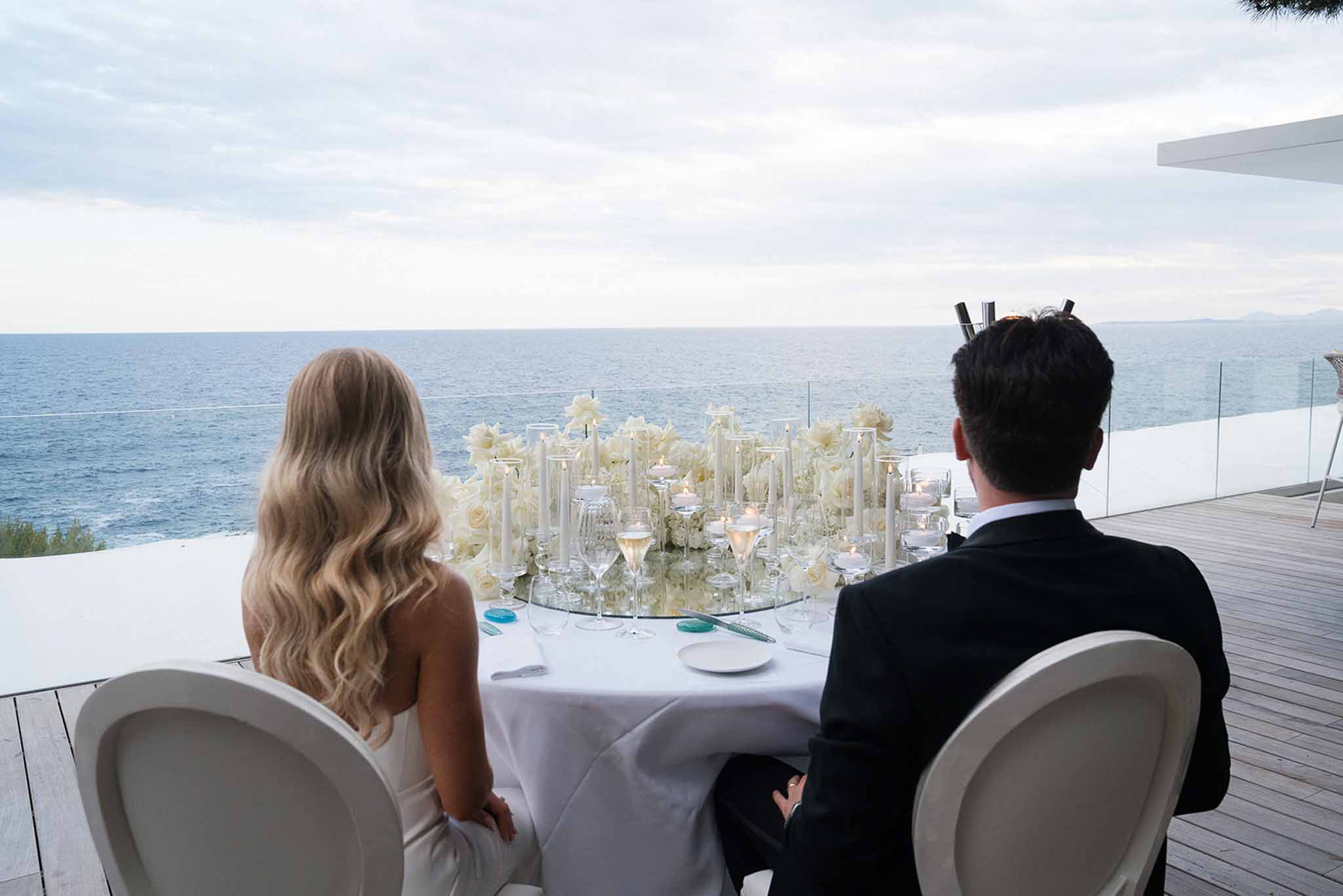 Bride and groom seated at sweetheart table on ocean-facing terrace with white rose centerpiece and taper candles