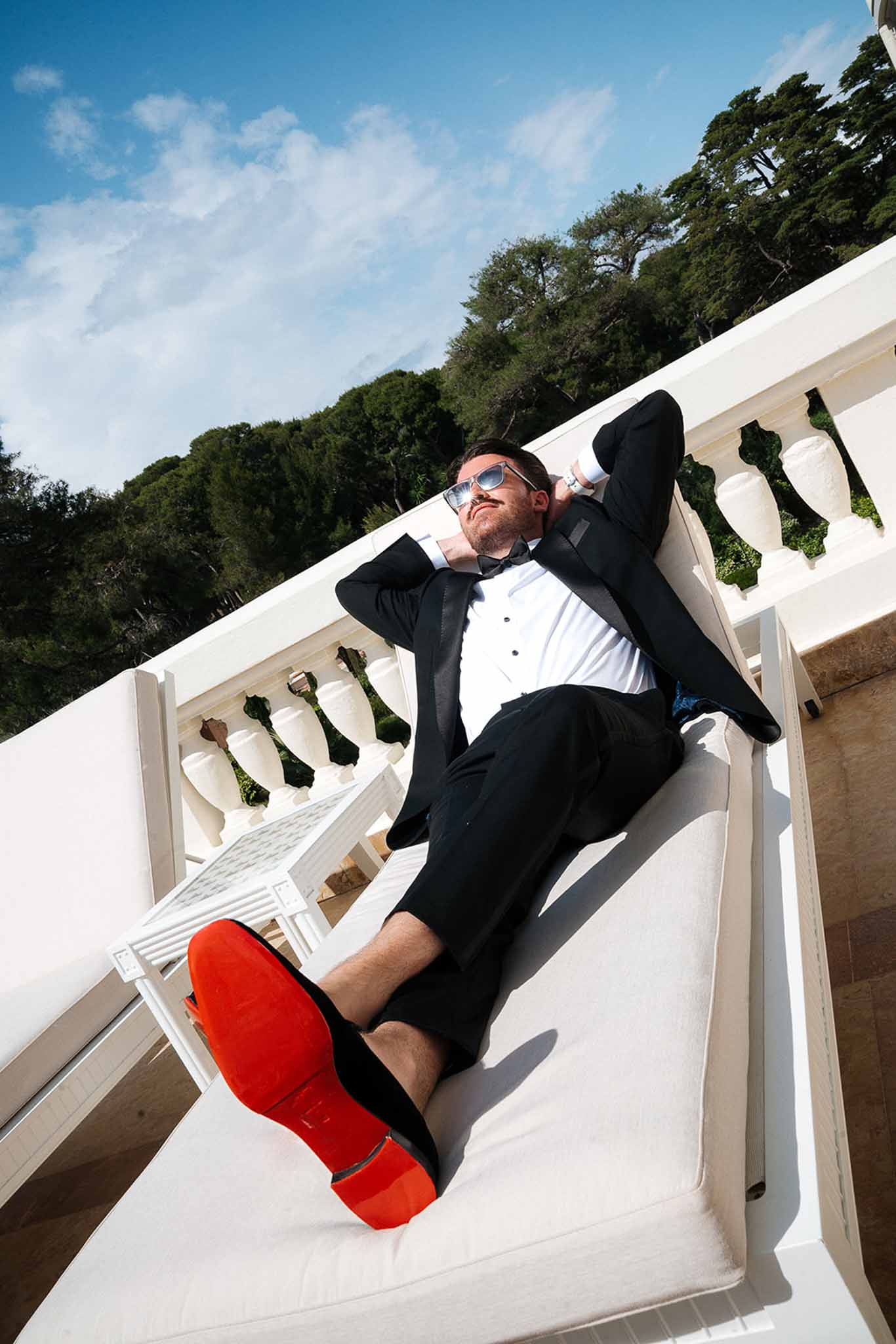 Groom in black tuxedo and mirrored aviators reclining on white terrace lounger with red-soled shoes and balustrade