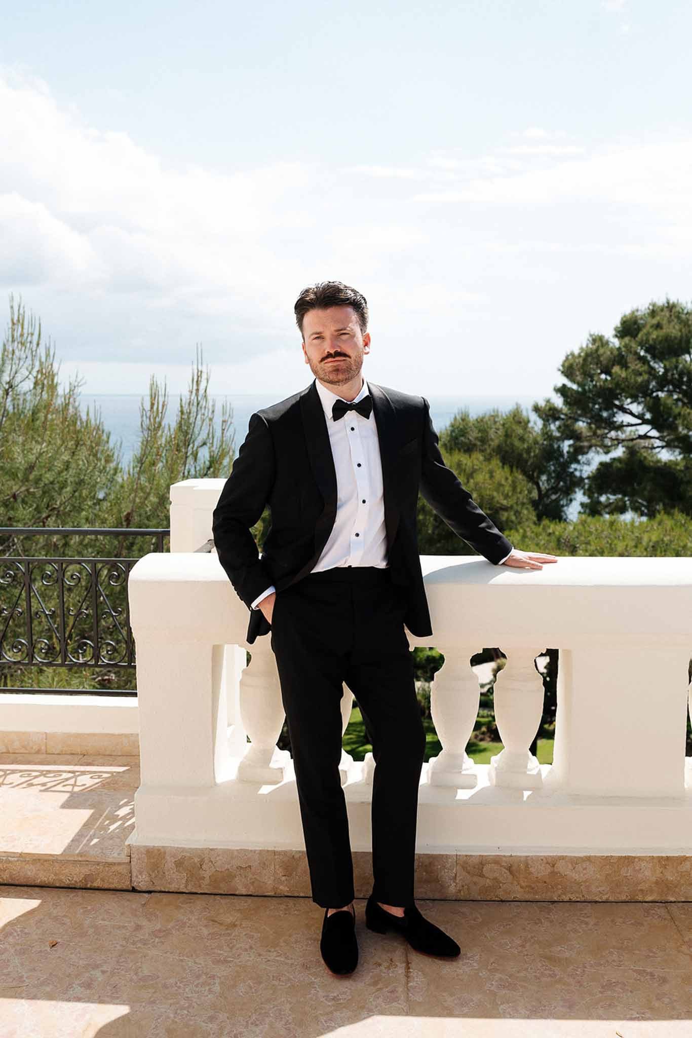Groom in black tuxedo leaning on white balustrade with coastal sea view behind