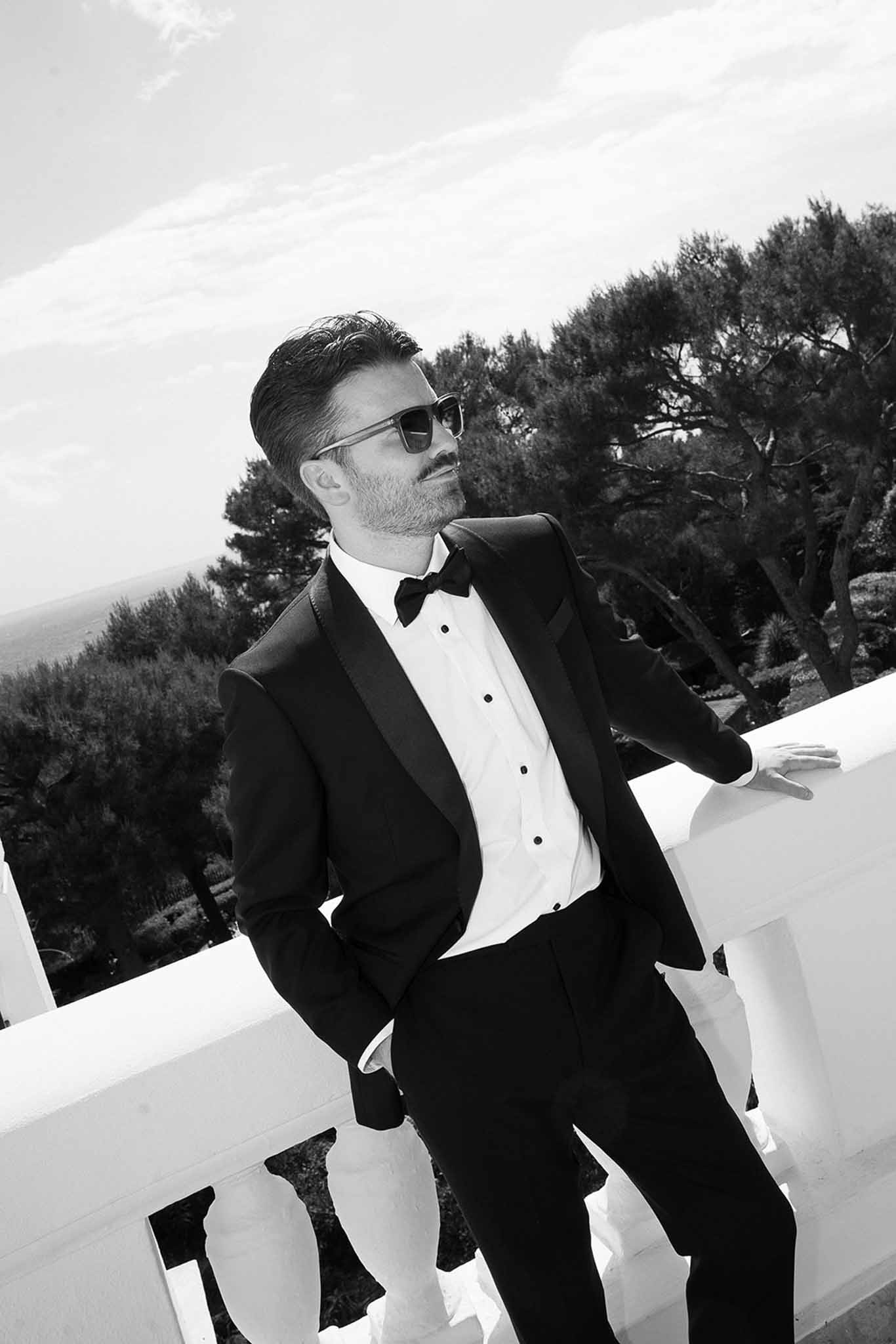 Black and white groom in black tuxedo with sunglasses leaning against white balustrade on outdoor terrace
