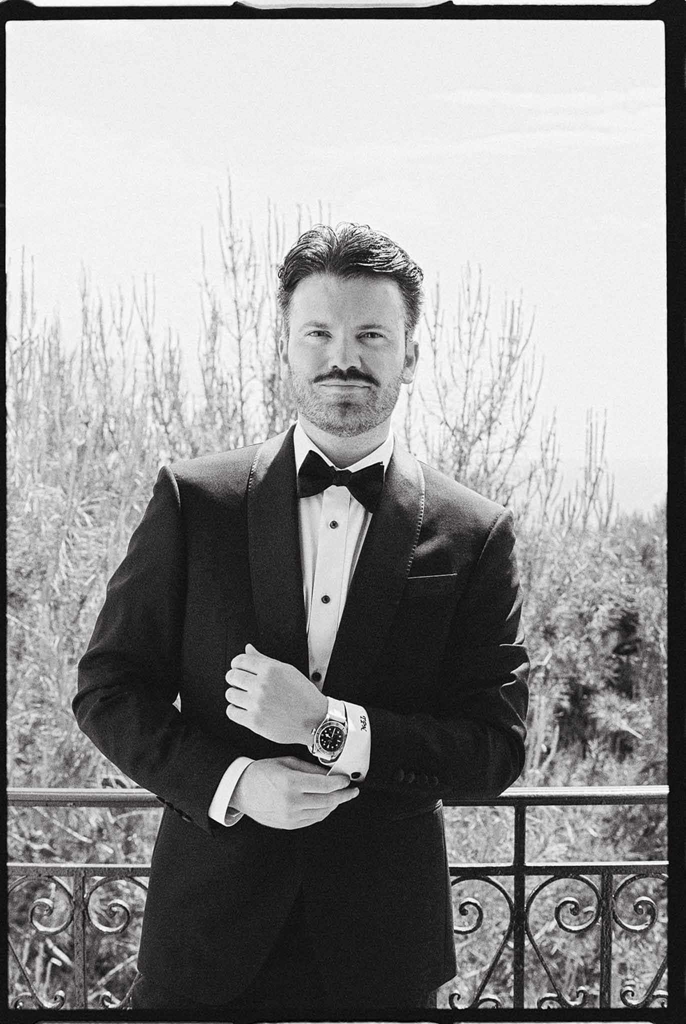 Black-and-white film portrait of groom in shawl-lapel tuxedo adjusting watch on outdoor terrace