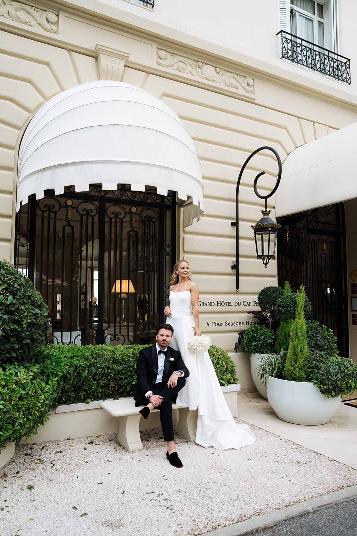 Groom sits on bench as bride stands with peony bouquet at Grand-Hotel du Cap-Ferrat wrought-iron entrance