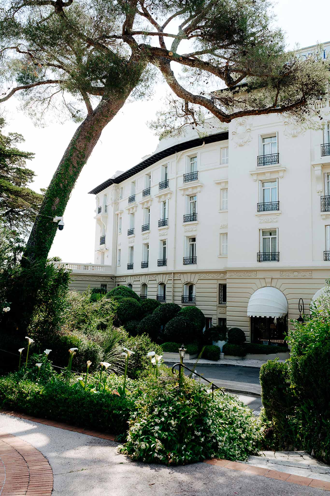 White Belle Epoque hotel with curved facade, iron balconies, and box topiary garden approach