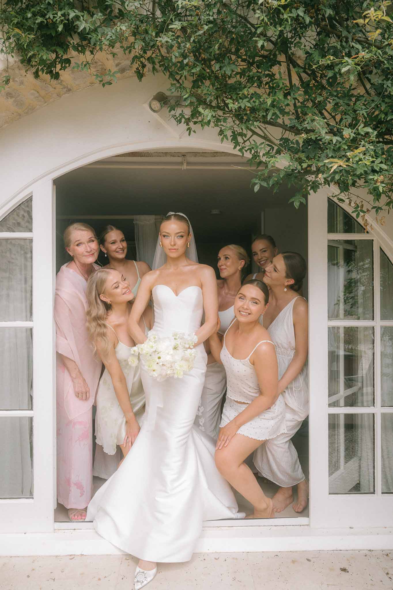 Bride in strapless satin gown with cathedral veil and six companions in robes posed in arched stone doorway