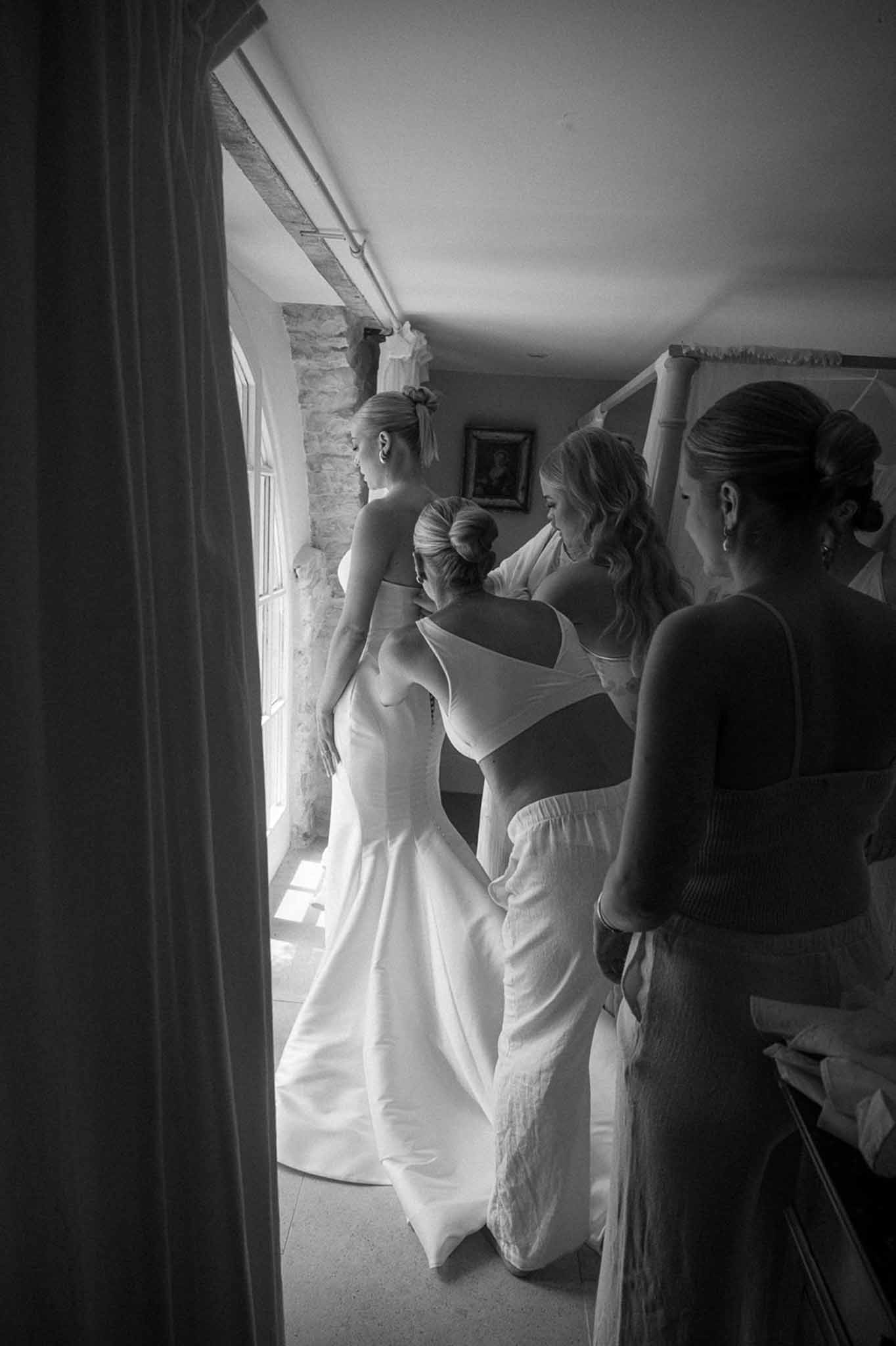 Black and white candid of bride being helped into dress by two attendants in stone-walled room