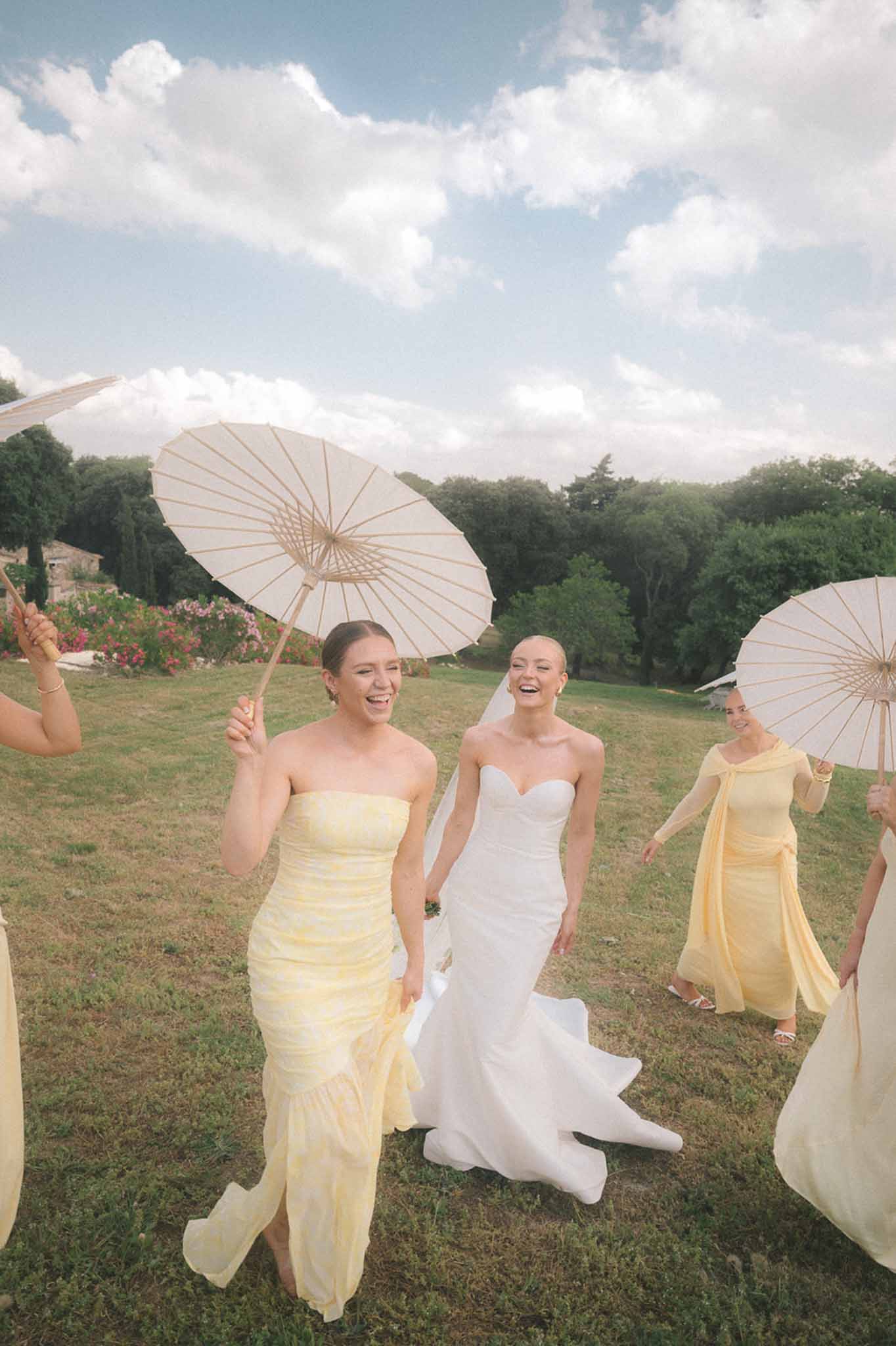 Bride and bridesmaids in pale yellow gowns walking across lawn each carrying white paper parasols