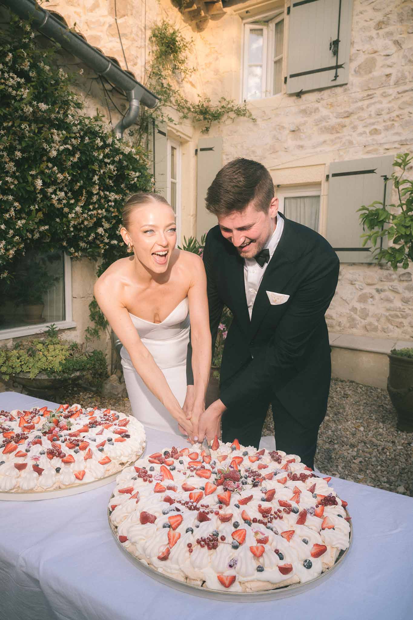 Laughing couple cutting pavlovas topped with fresh berries outdoors at stone-facade venue