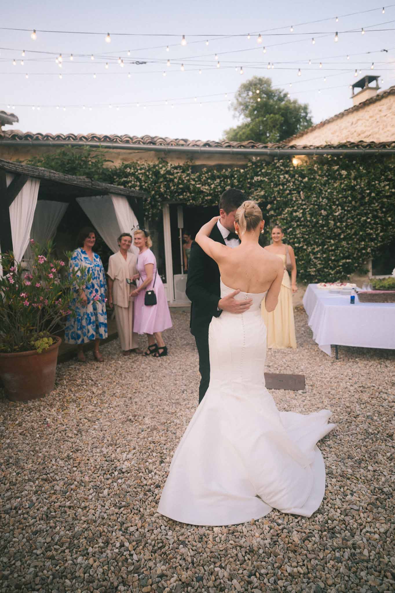 Couple sharing first dance on gravel courtyard at dusk under string lights with guests watching nearby