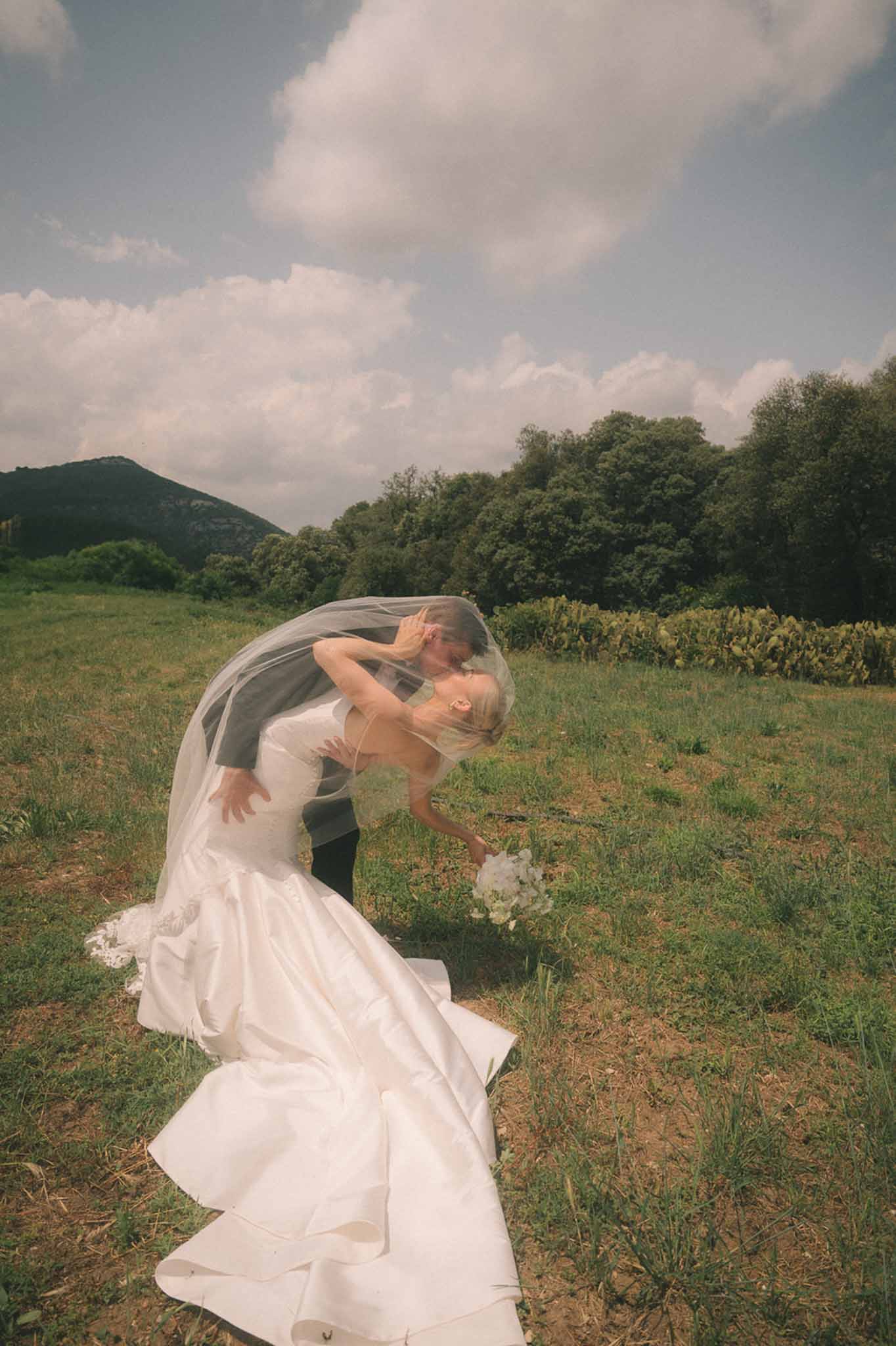 Groom dipping bride into a kiss as cathedral-length veil billows over them in open field with mountain backdrop