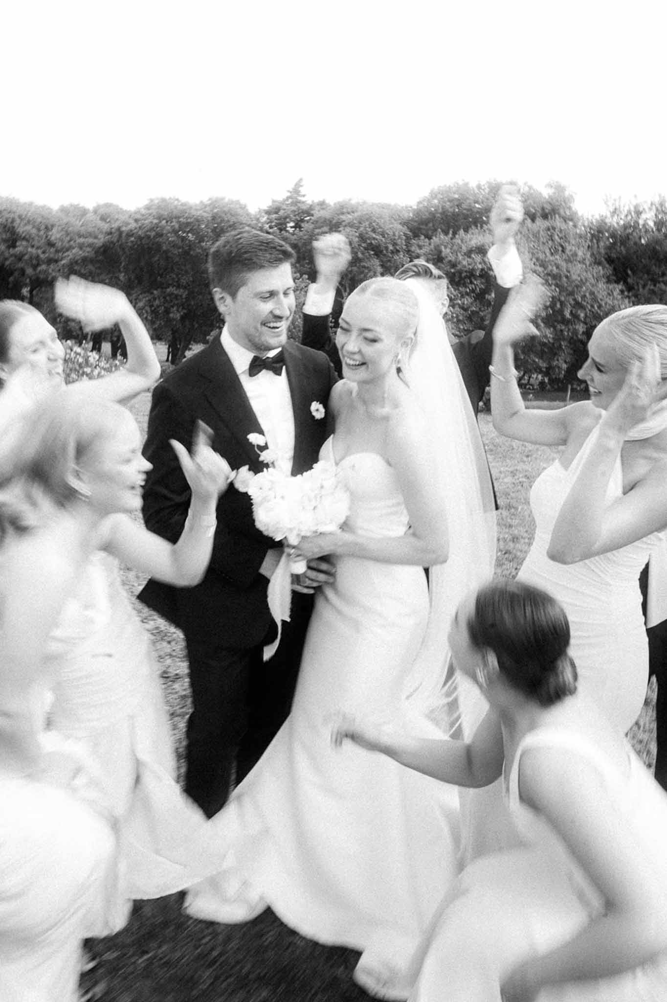 Black and white bridal party cheering around couple with motion blur under trees