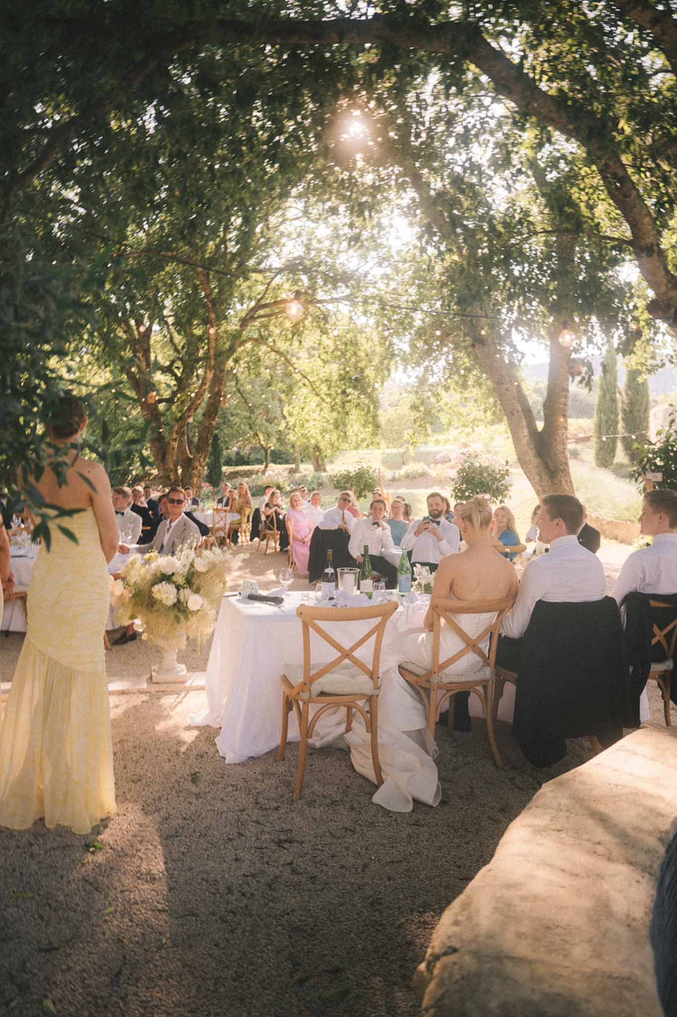 Guest delivering a speech at an outdoor reception under mature trees with string lights and seated guests at round tables