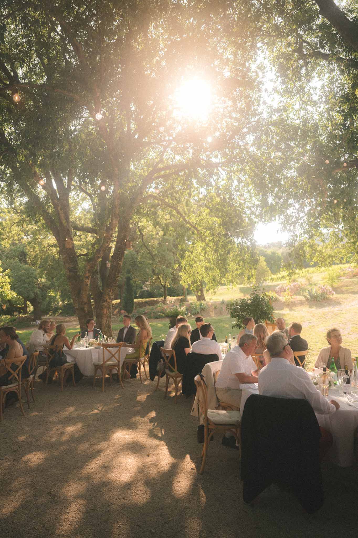 Outdoor wedding reception dinner under mature trees with string lights, guests seated at round tables on gravel during gol...