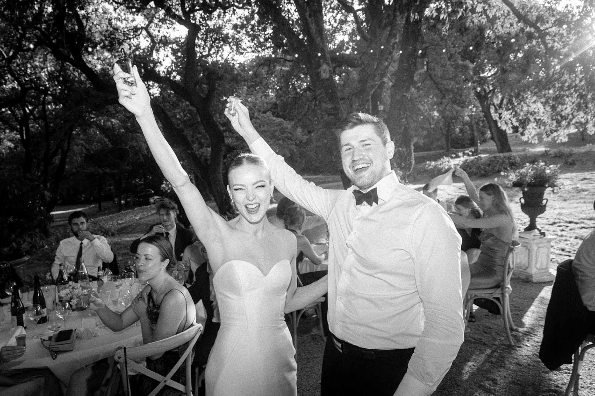 Black and white candid of bride raising shot glass while laughing beside groom under fairy-lit trees