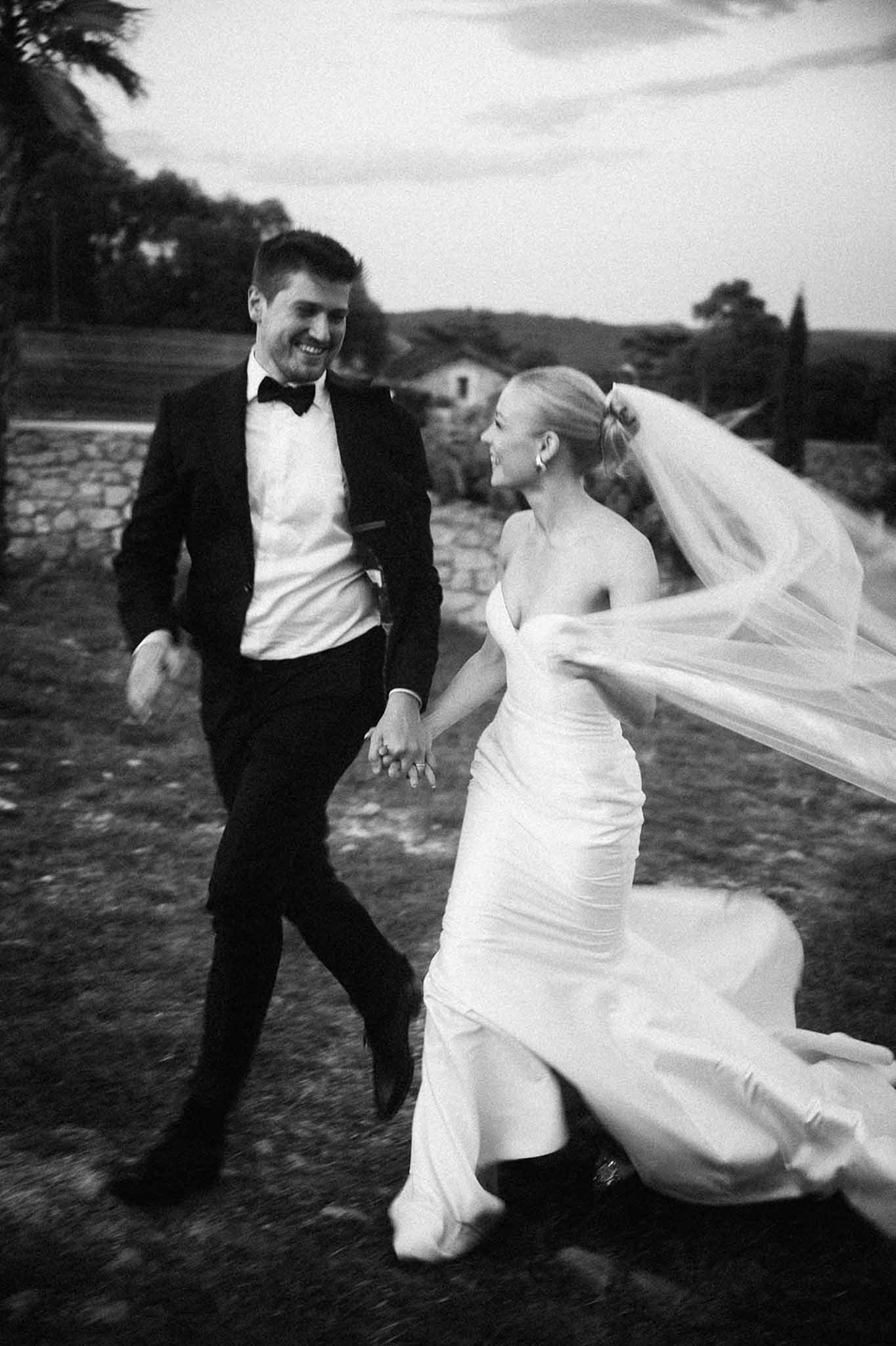 Black and white photo of bride and groom running and laughing hand in hand with veil billowing at dusk