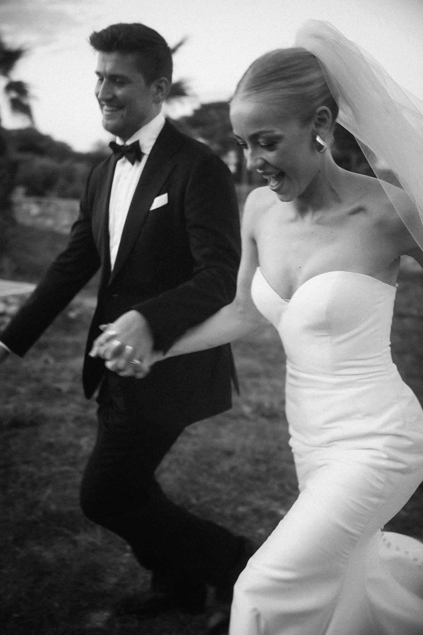 Black and white photo of bride and groom running and laughing while holding hands outdoors