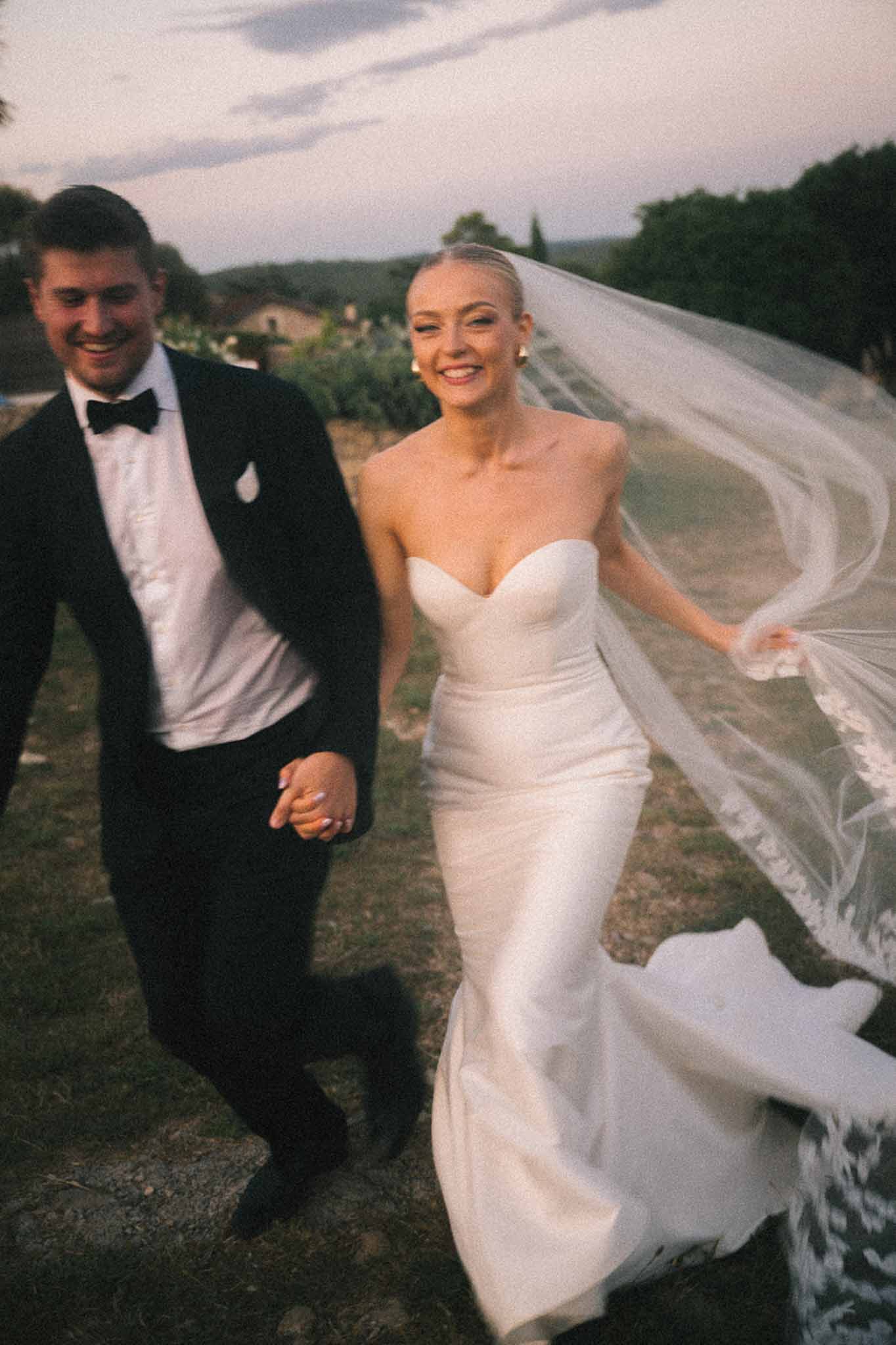 Couple walking with lace-trimmed veil billowing in dusk light with vineyard hills in background