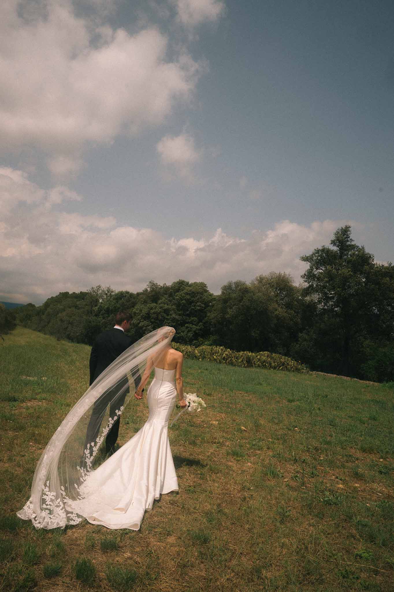 Couple walks hand-in-hand across hillside as wind catches lace-trimmed cathedral veil in film tones