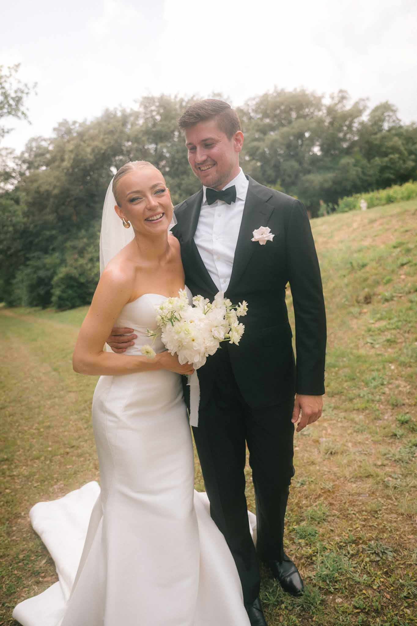 Bride in strapless white satin gown with groom in black tuxedo smiling together on grassy hillside