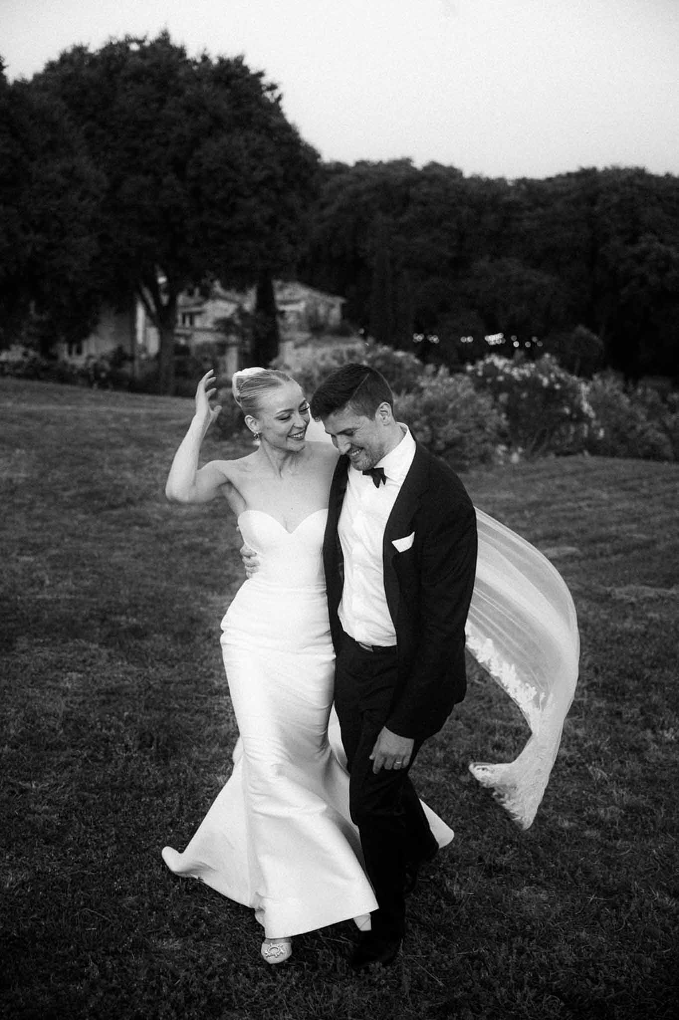 Black and white couple walking on lawn with lace-trimmed veil billowing, bride in mermaid gown laughing