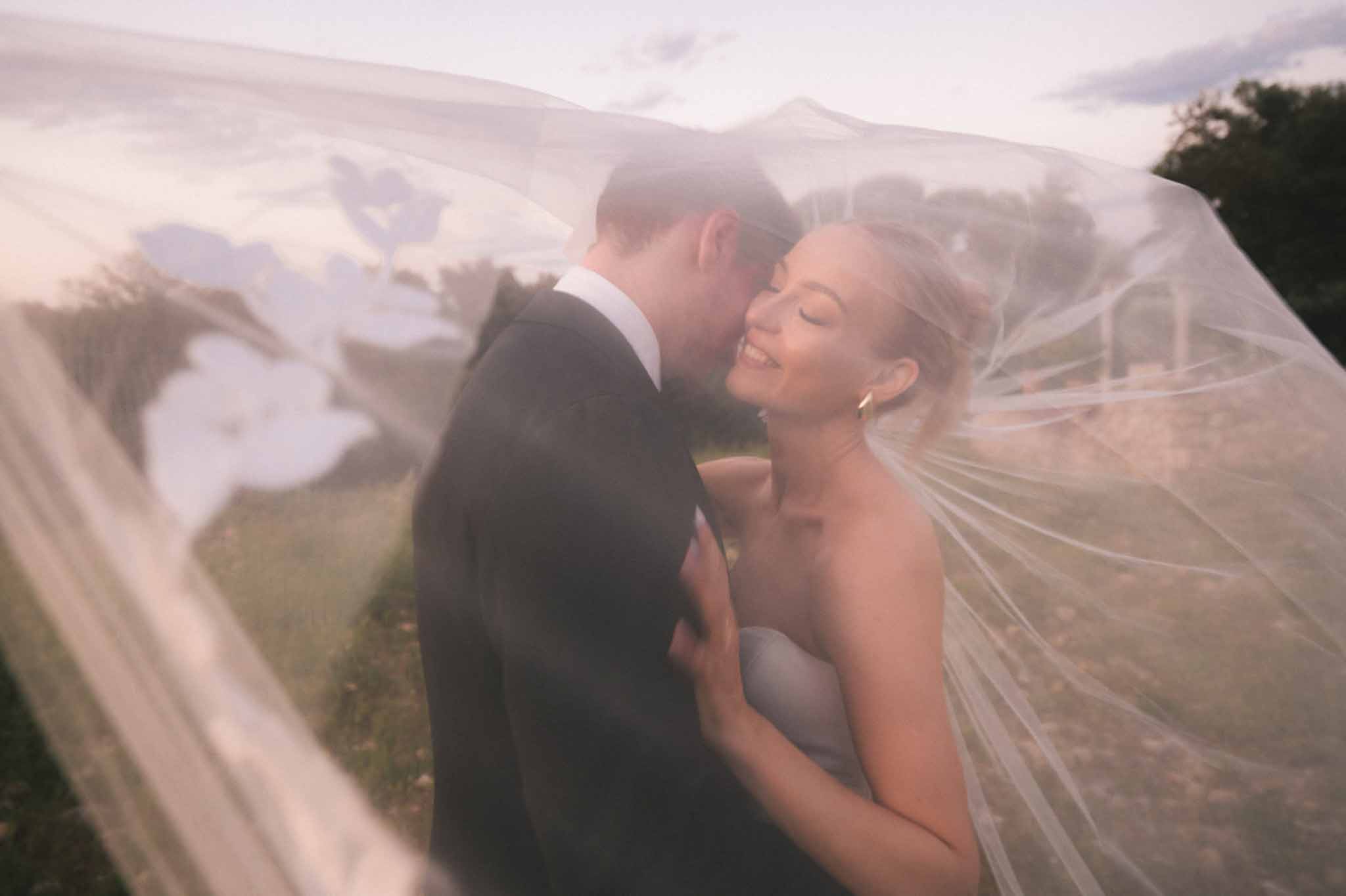 Groom kisses bride on the cheek at golden hour as her embroidered cathedral veil billows around them both