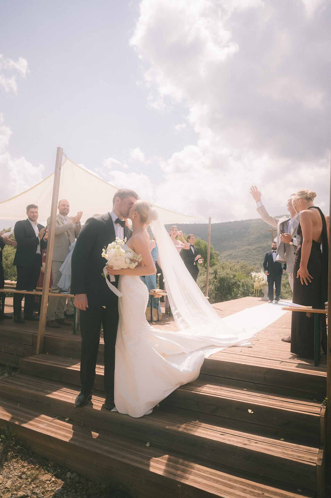 First kiss on hilltop wooden deck with cathedral veil on steps and cream shade sail canopy above