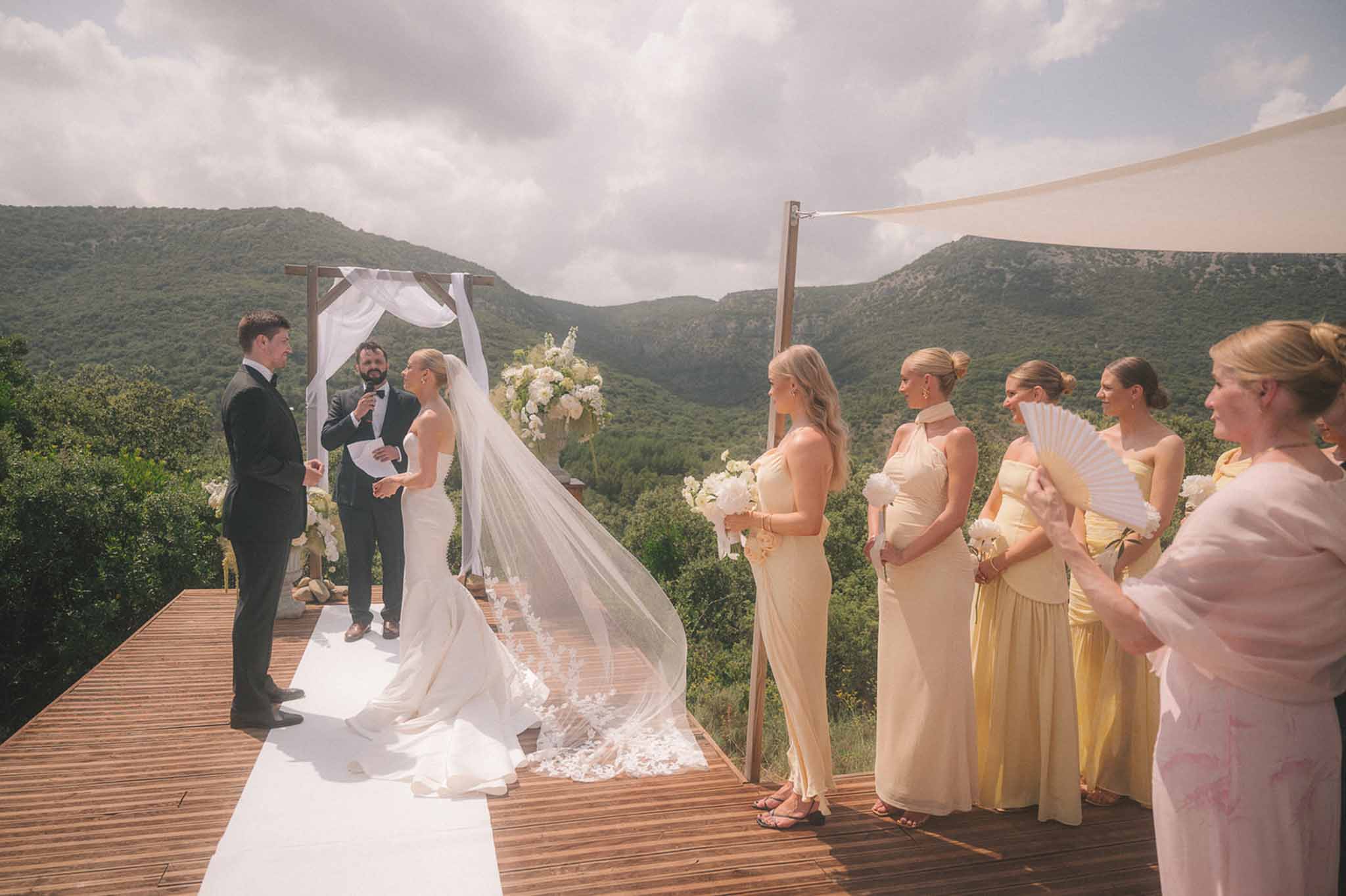Outdoor mountain ceremony with couple under draped wooden arch, bridesmaids in yellow gowns on elevated terrace