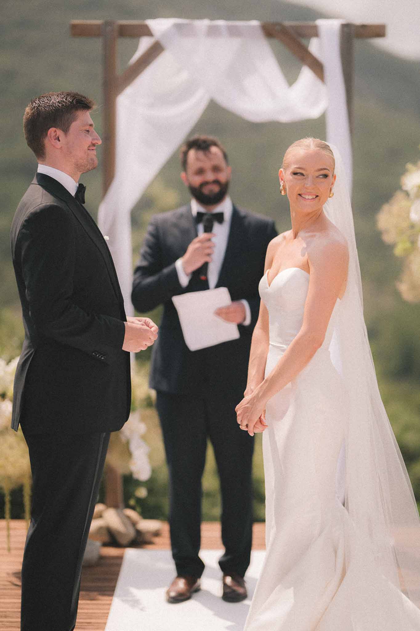 Smiling bride and groom holding hands at outdoor altar with draped wooden arch and mountain view