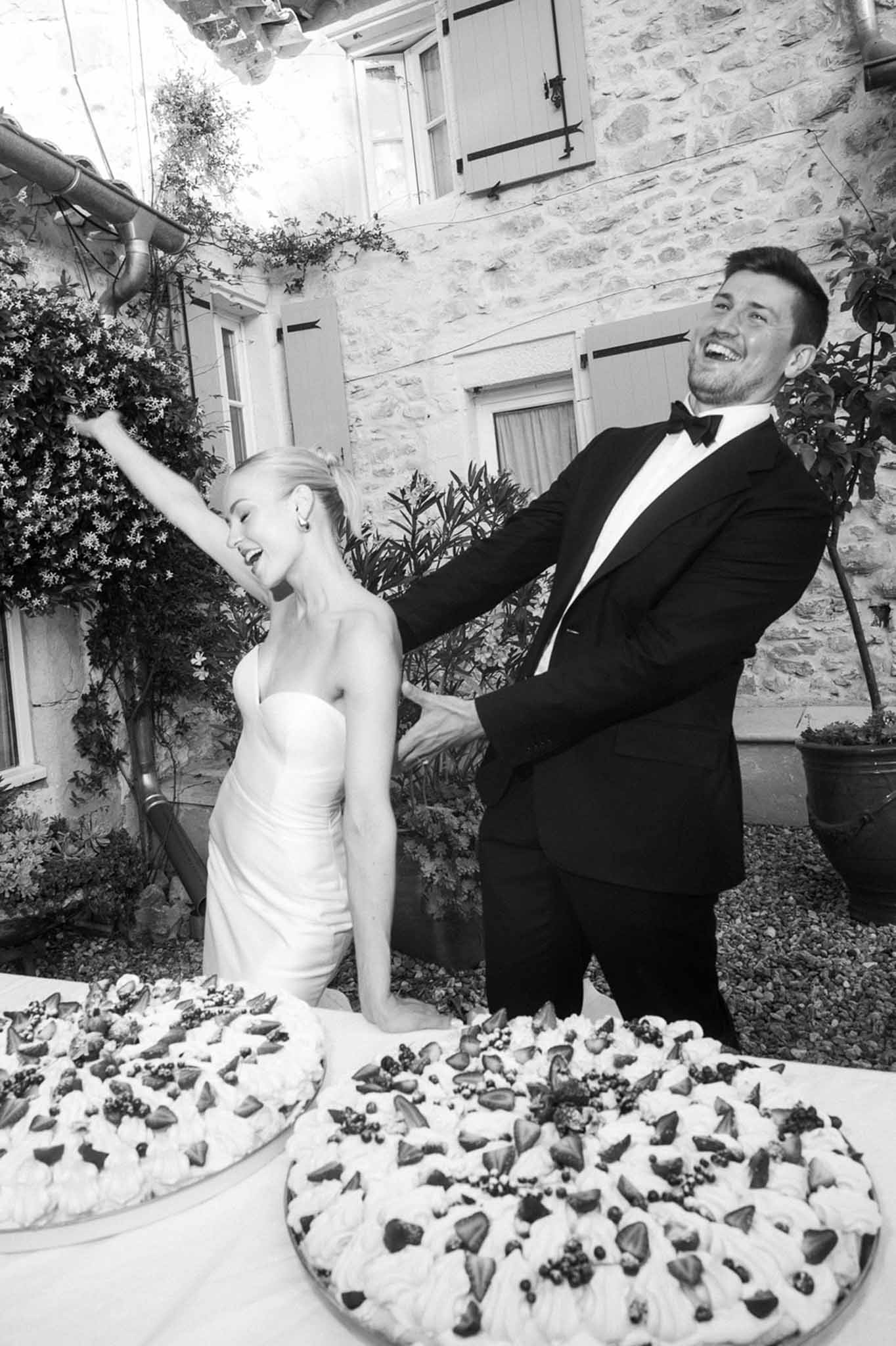 Black and white couple laughing behind two pavlova cakes with strawberries at outdoor reception by stone farmhouse