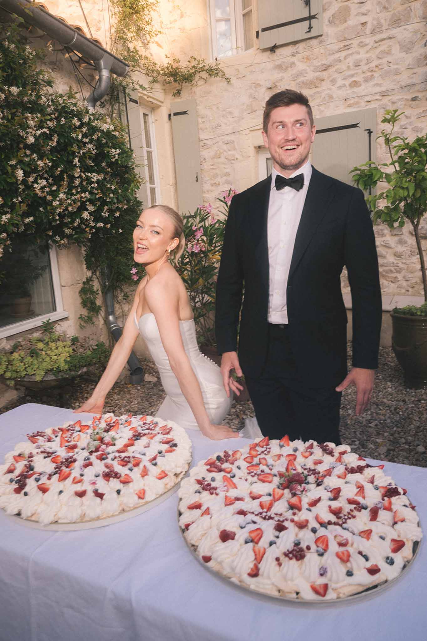 Bride and groom laughing beside two pavlova cakes in a stone courtyard at dusk