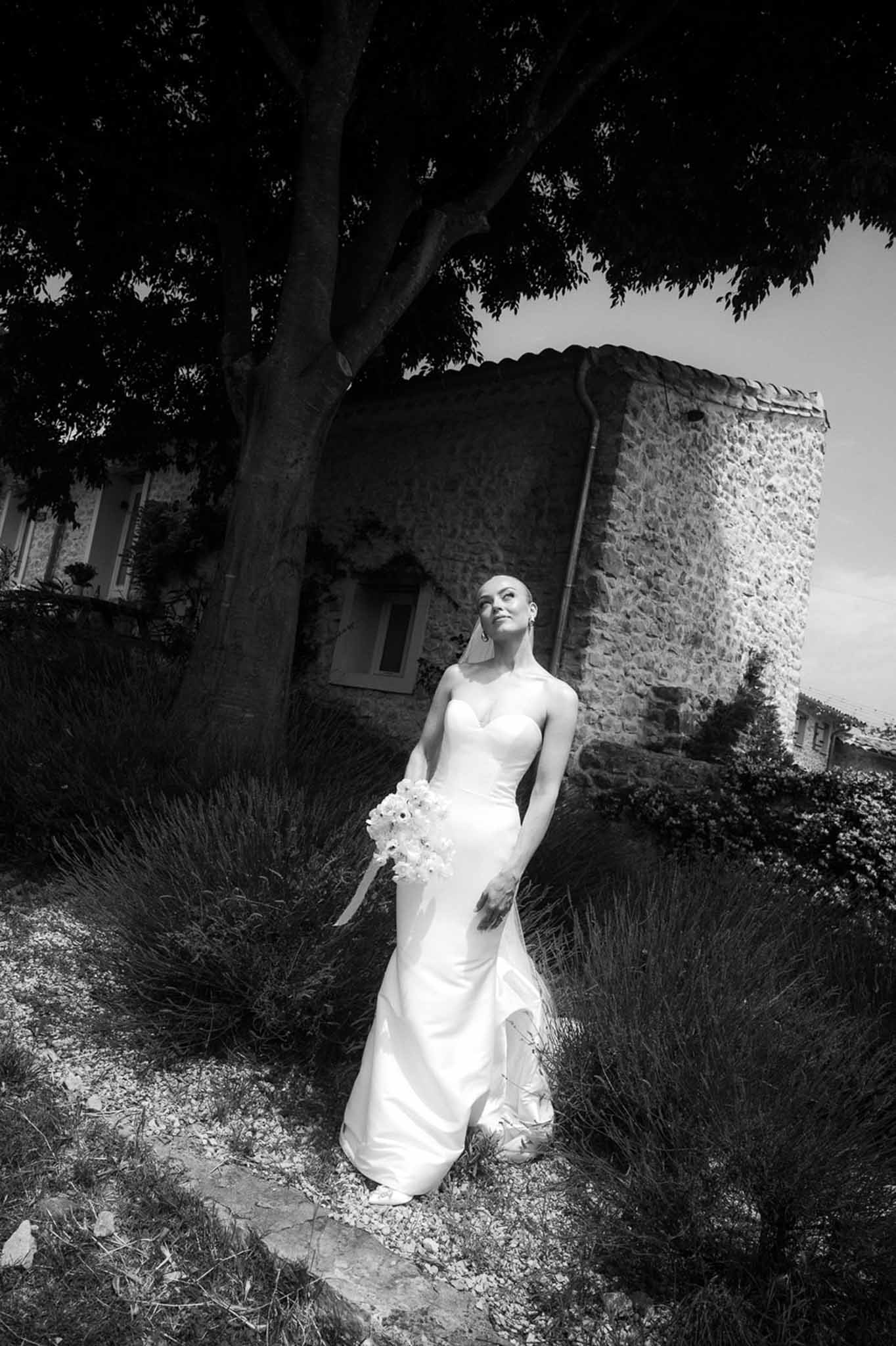 Black and white portrait of bride in strapless gown holding bouquet beneath tree with stone tower in background