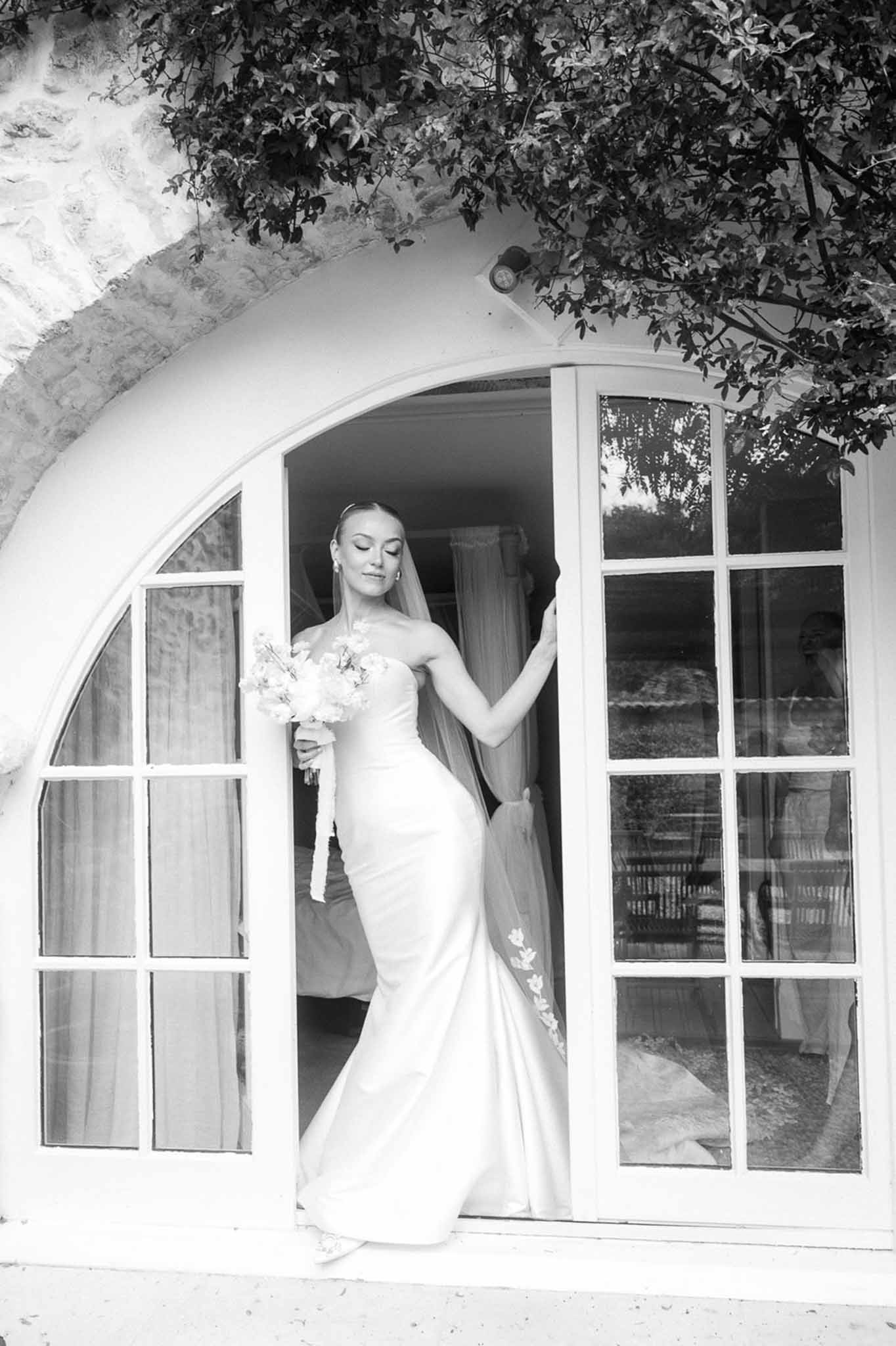 Black and white of bride stepping through stone arch in mermaid gown with floral-edged veil and bouquet