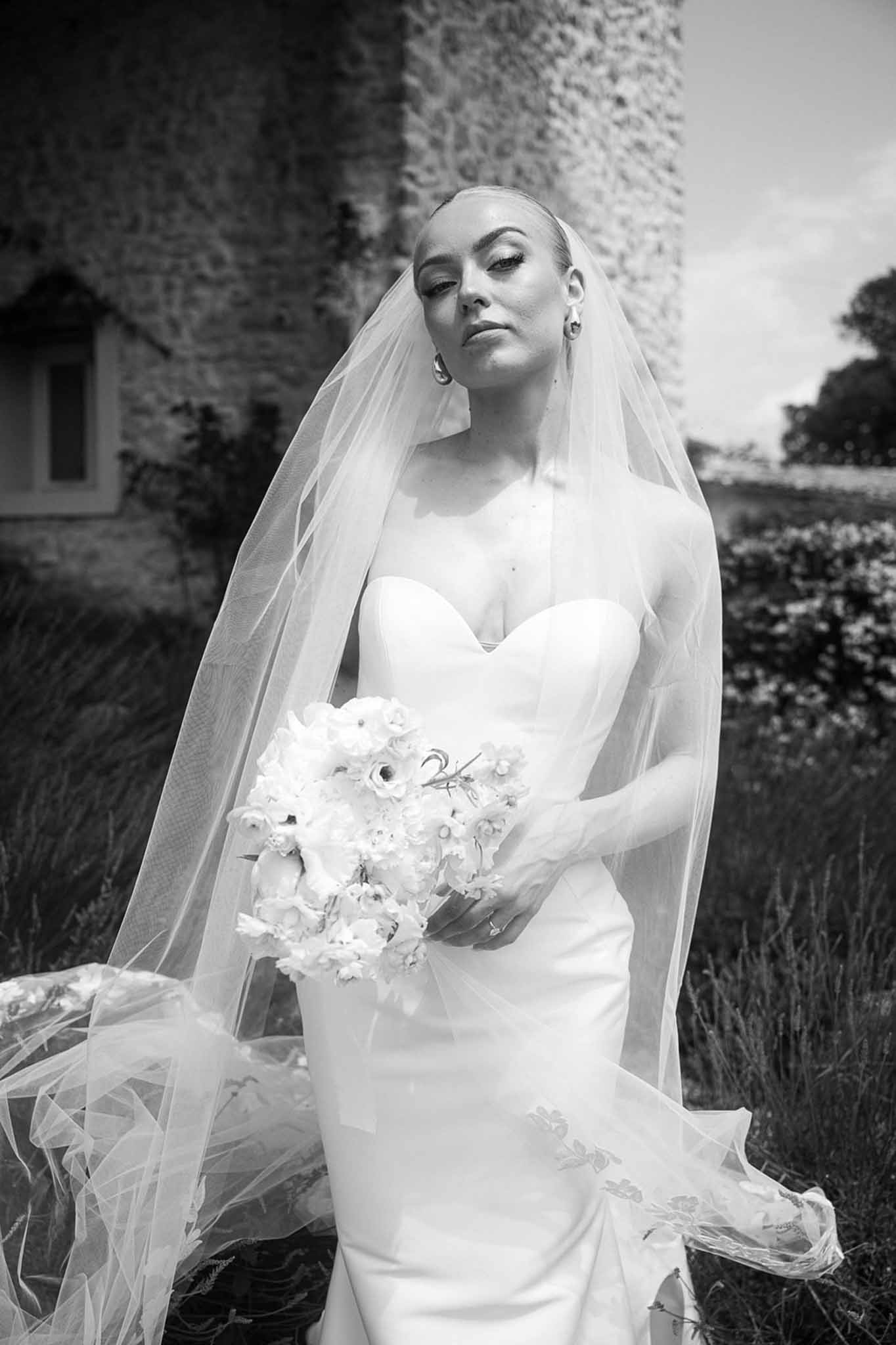 Black and white bridal portrait of bride in strapless gown with cathedral veil blowing in wind outside stone chateau