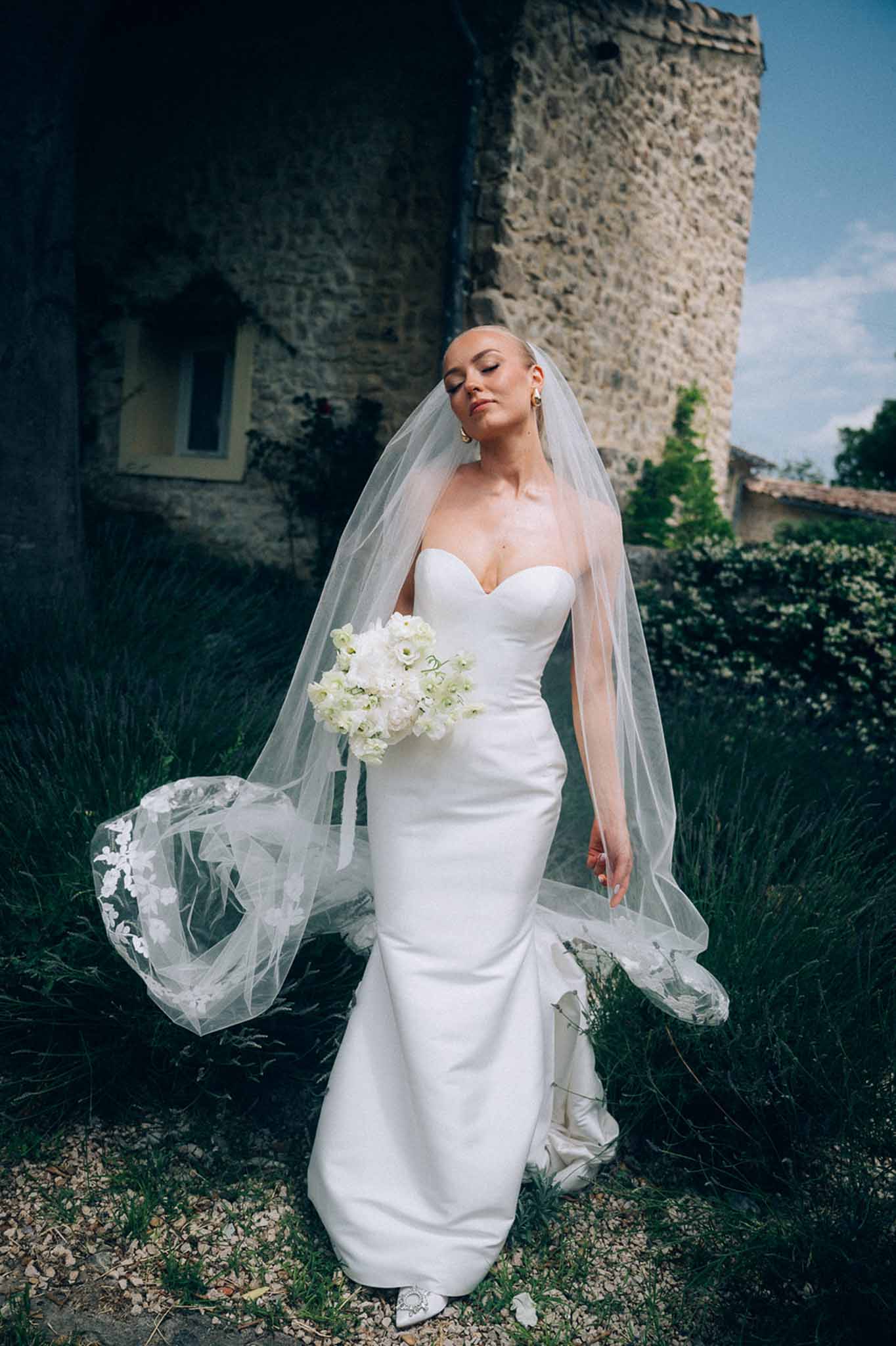 Bride in strapless ivory satin gown with cathedral-length lace veil and white peony bouquet beside stone chateau