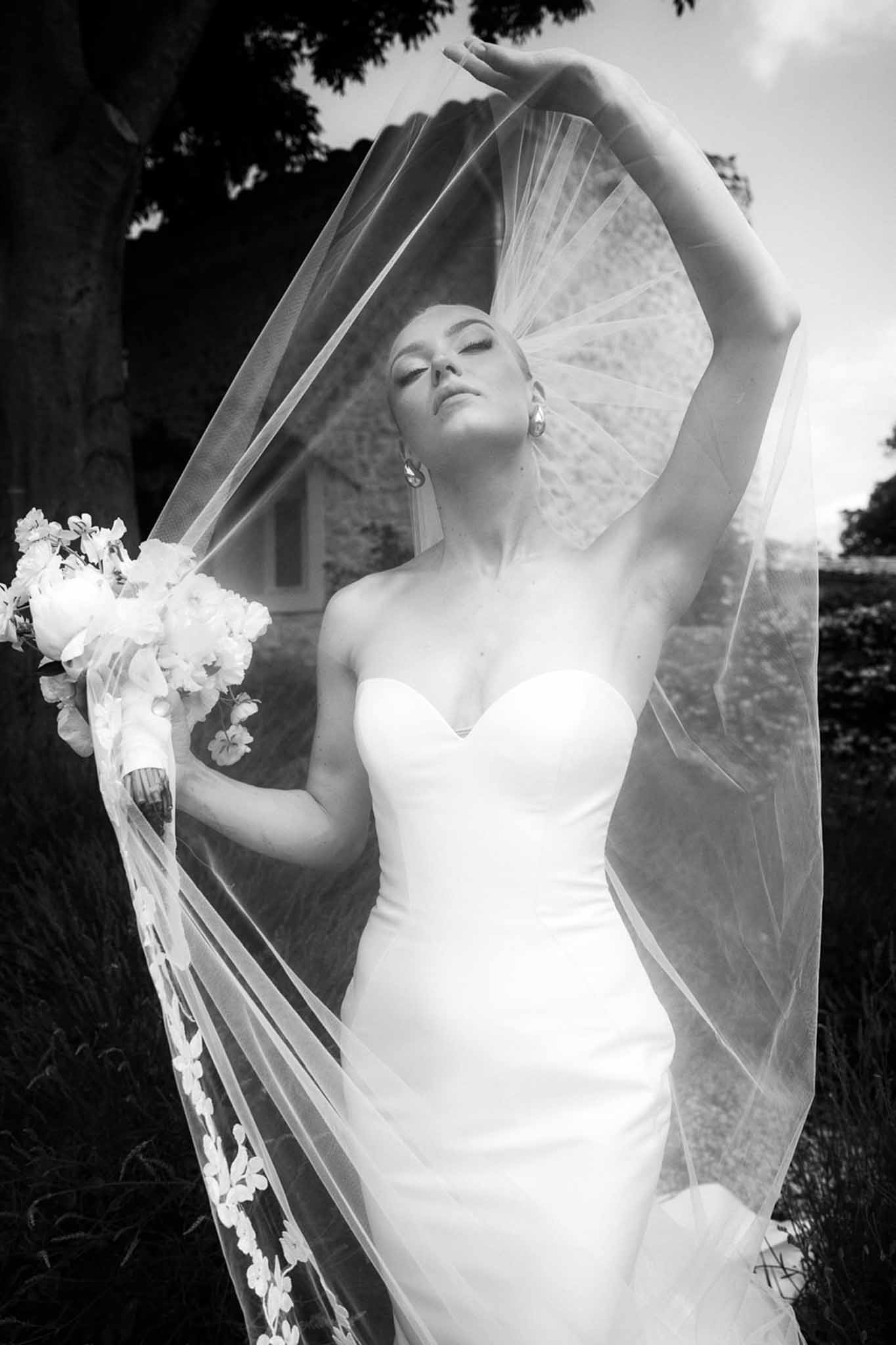 Black and white portrait of bride lifting embroidered veil overhead in strapless gown with peony bouquet