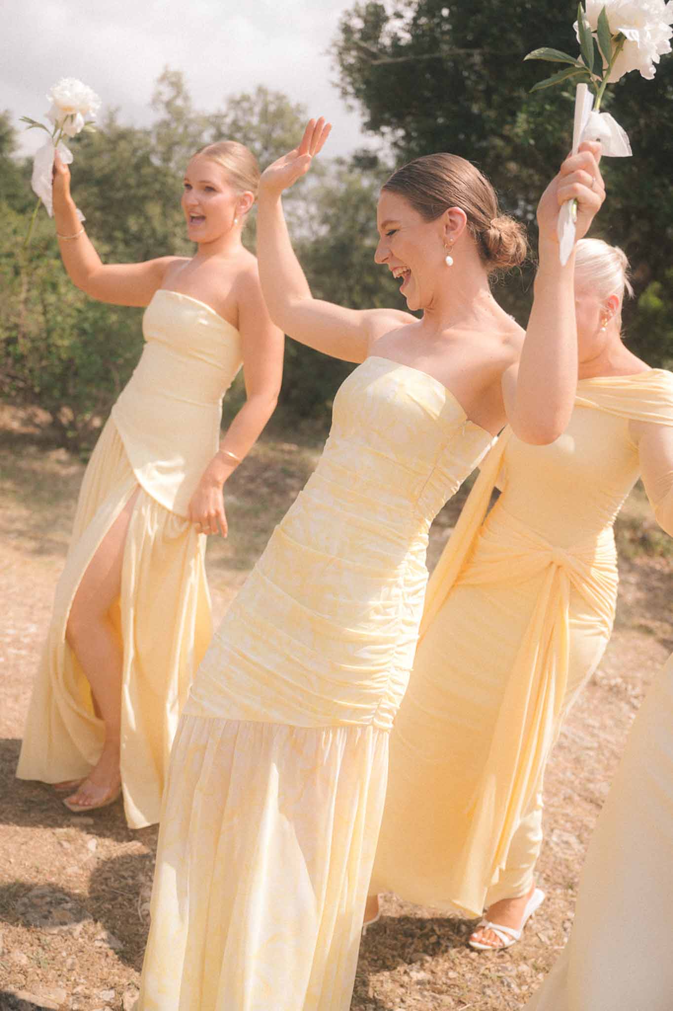 Three bridesmaids in pale yellow strapless dresses raising peonies and celebrating on dirt path