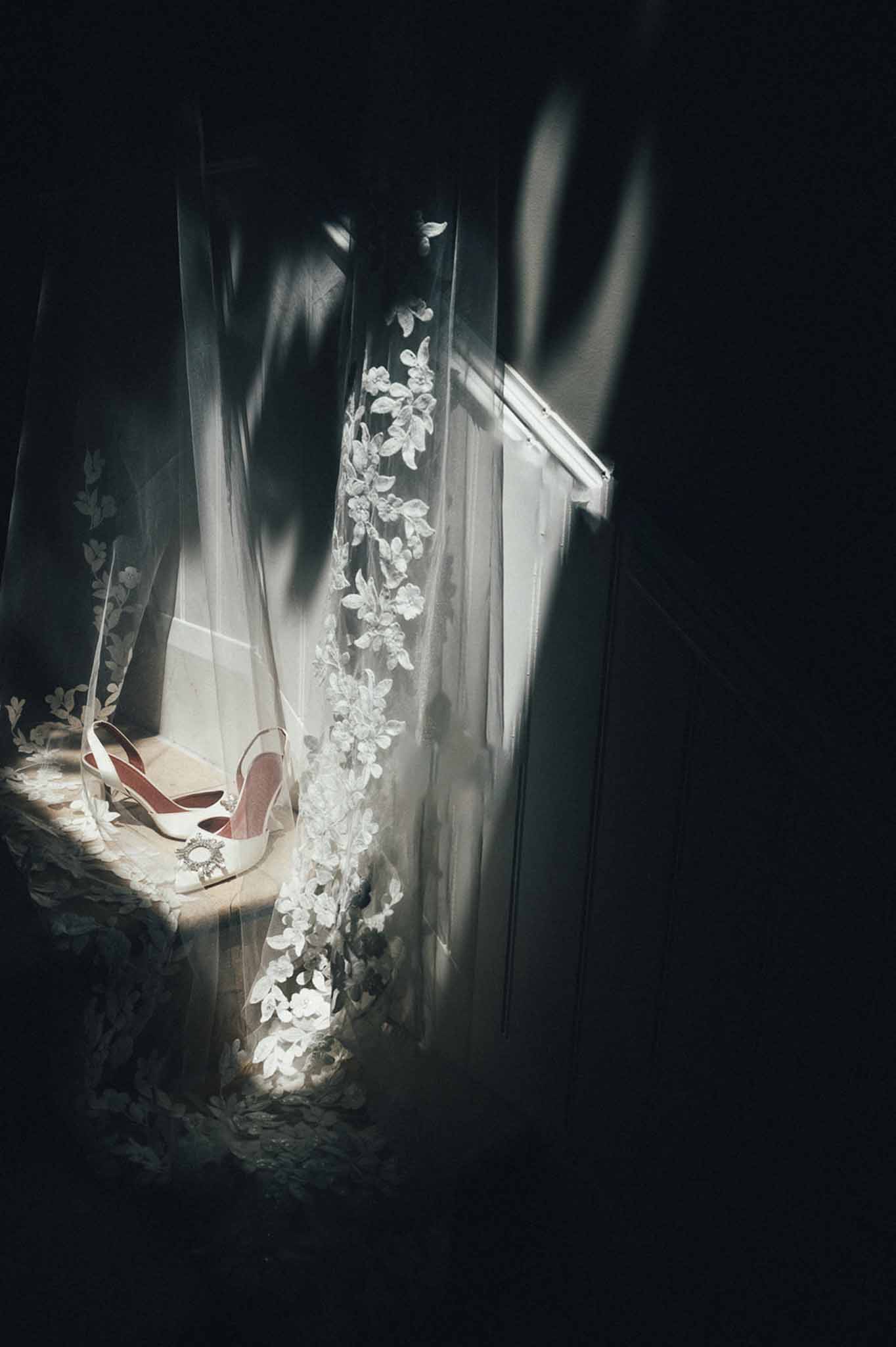 White satin bridal heels with jewelled buckle and floral lace veil on window ledge in dramatic natural light