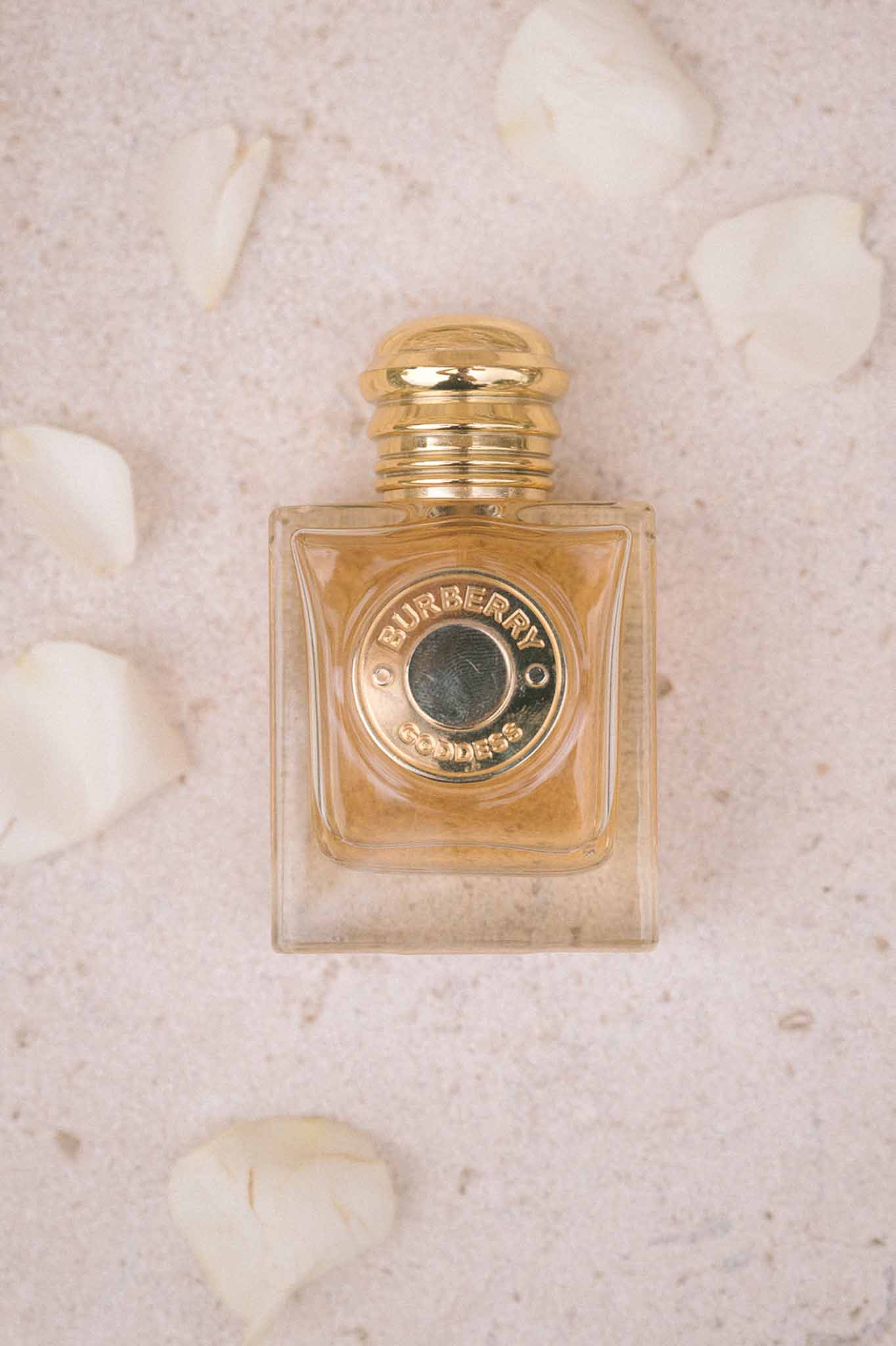 Burberry Goddess perfume bottle on blush stone surface surrounded by scattered ivory flower petals