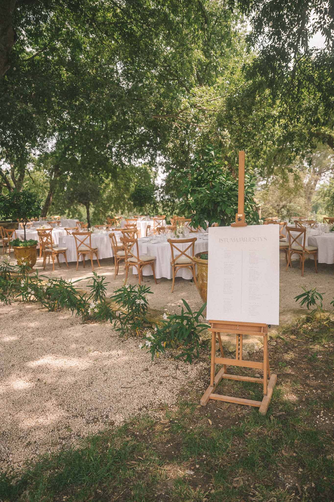 Wooden easel seating chart before cross-back chairs and long tables under mature trees on gravel garden