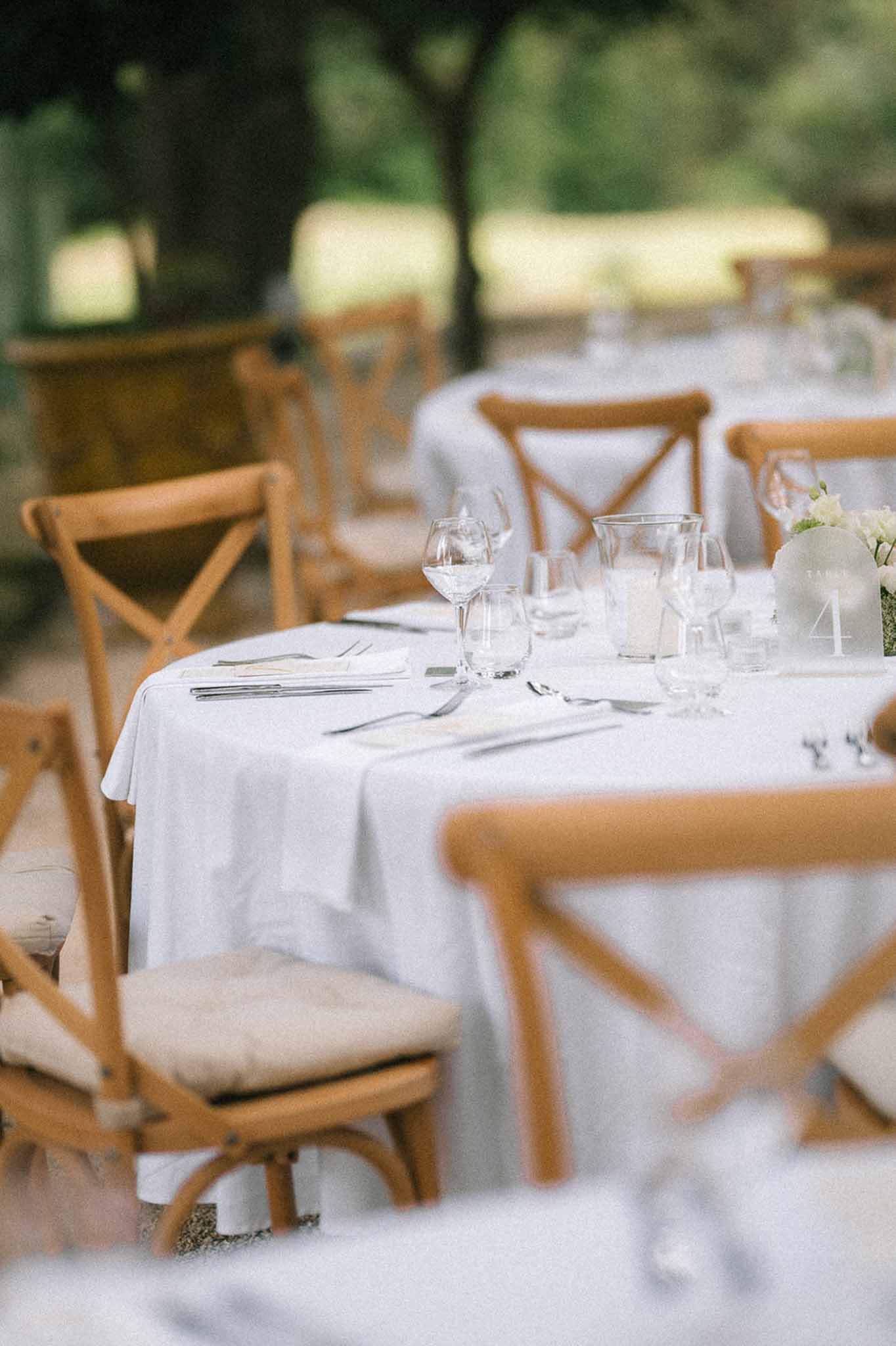 White linen reception table with cross-back chairs, silver cutlery, and frosted glass table number four in garden