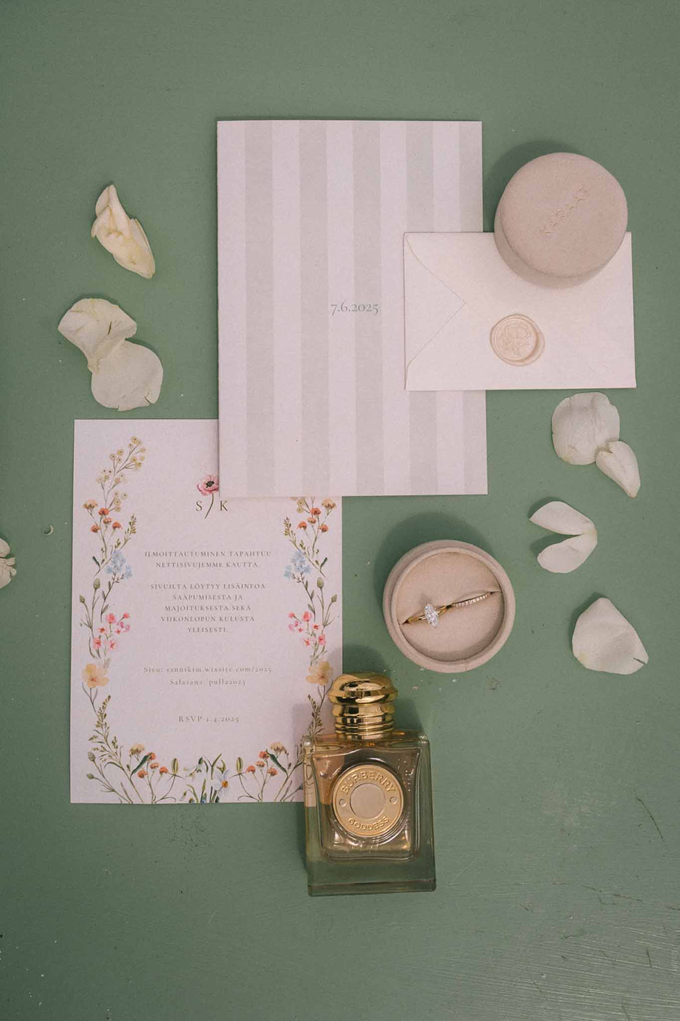Wedding stationery flat lay with wildflower invitations, velvet ring box, Burberry perfume, and scattered petals on sage s...