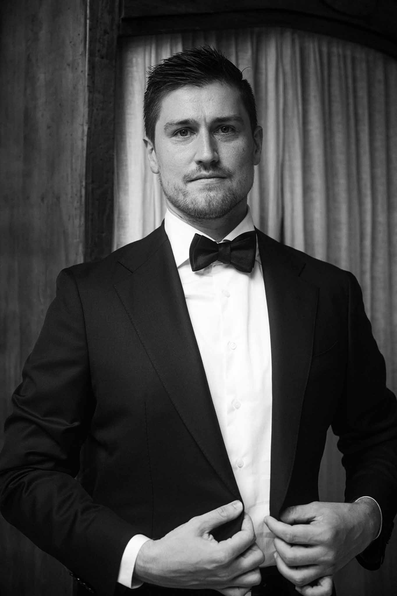 Black and white groom adjusting tuxedo jacket with bow tie looking directly at camera
