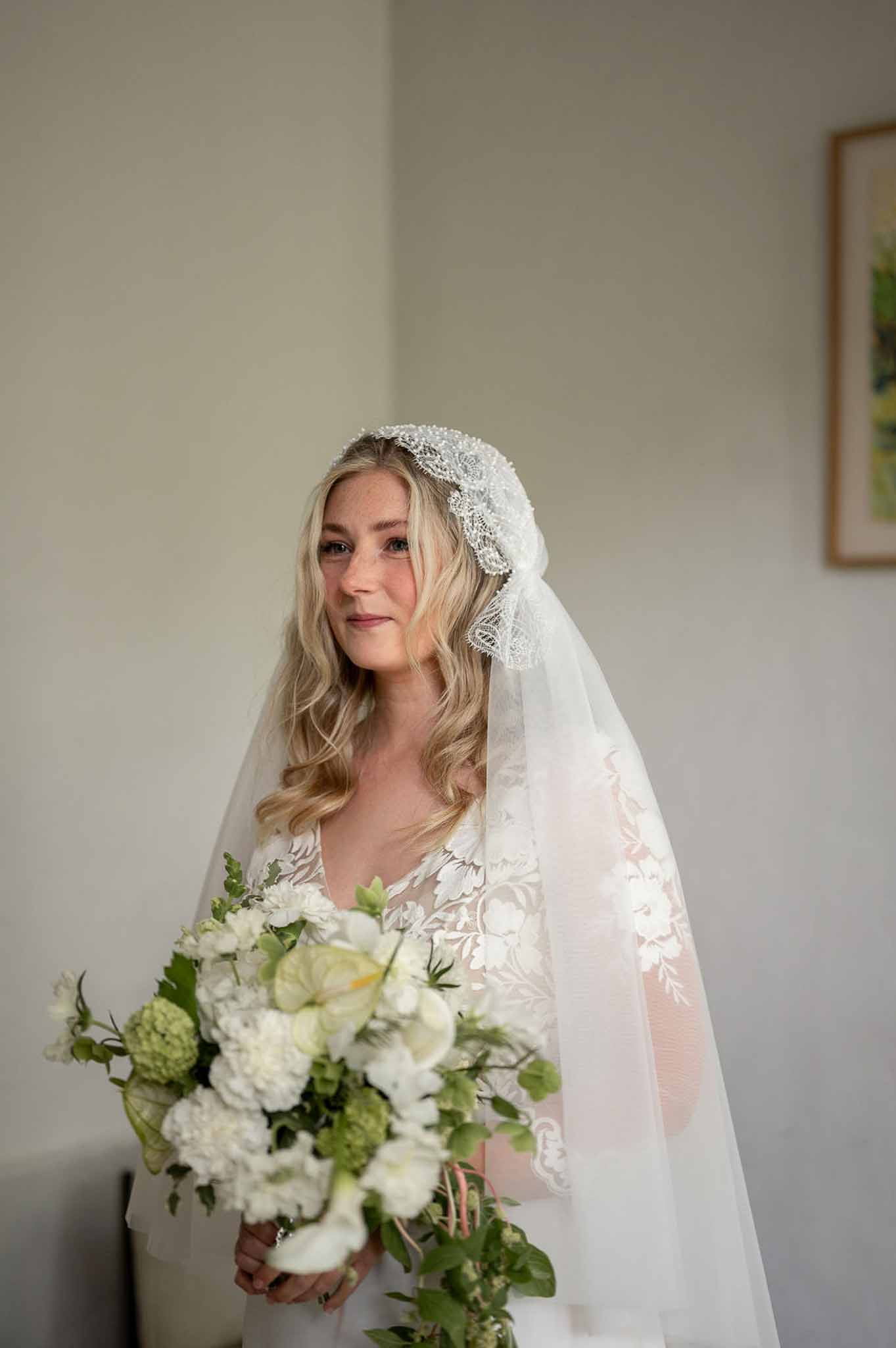 Bride in white lace gown and vintage Juliet-cap veil holding a loose white and green bouquet against a grey wall