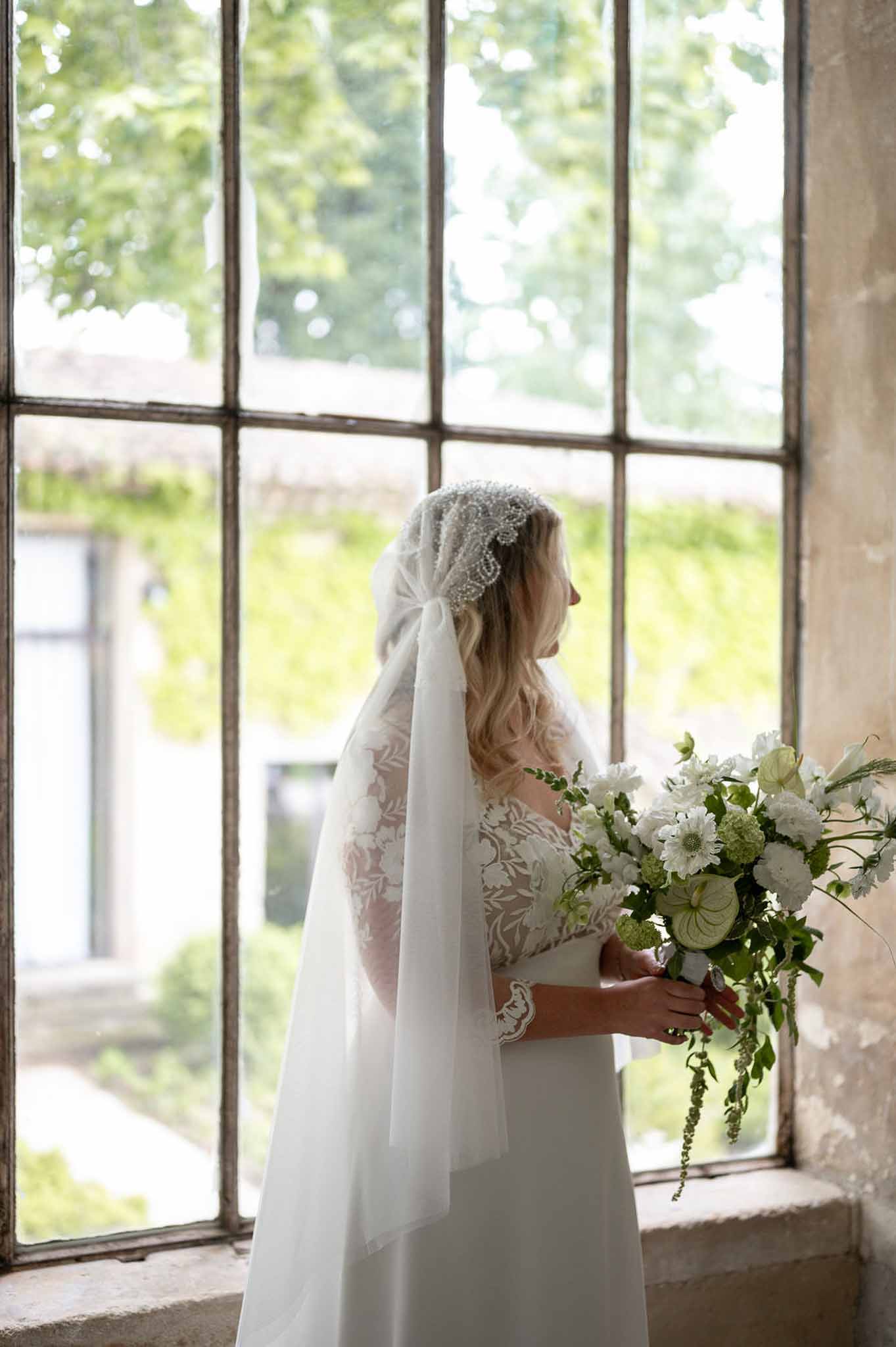 Bride in ivory lace gown and Juliet cap veil holding green viburnum and white scabiosa bouquet at industrial window