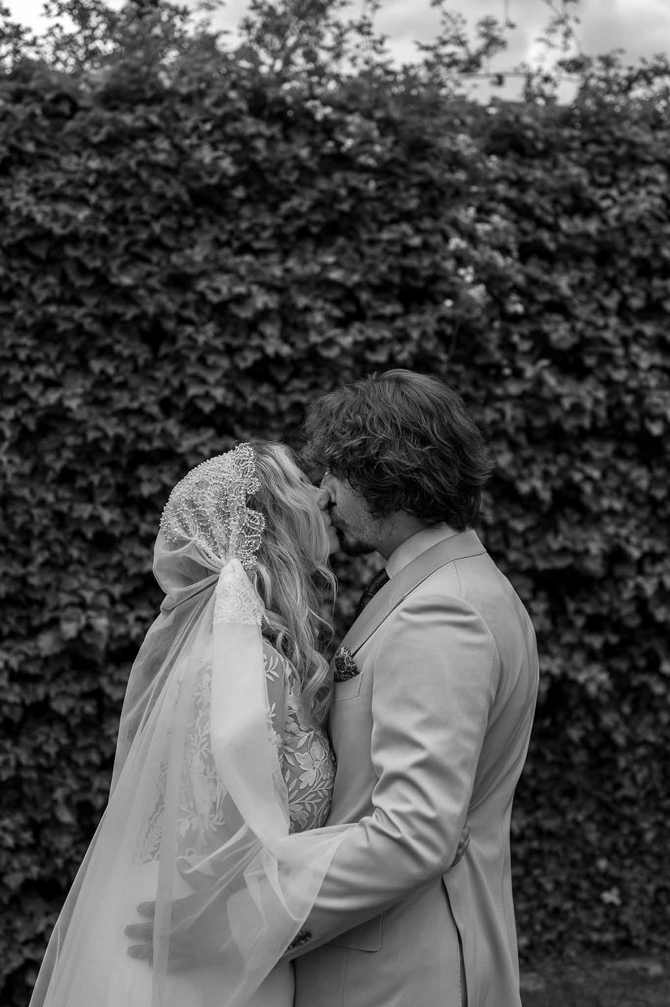 Bride and groom kissing in front of an ivy-covered wall with cathedral-length lace-trimmed veil in black and white