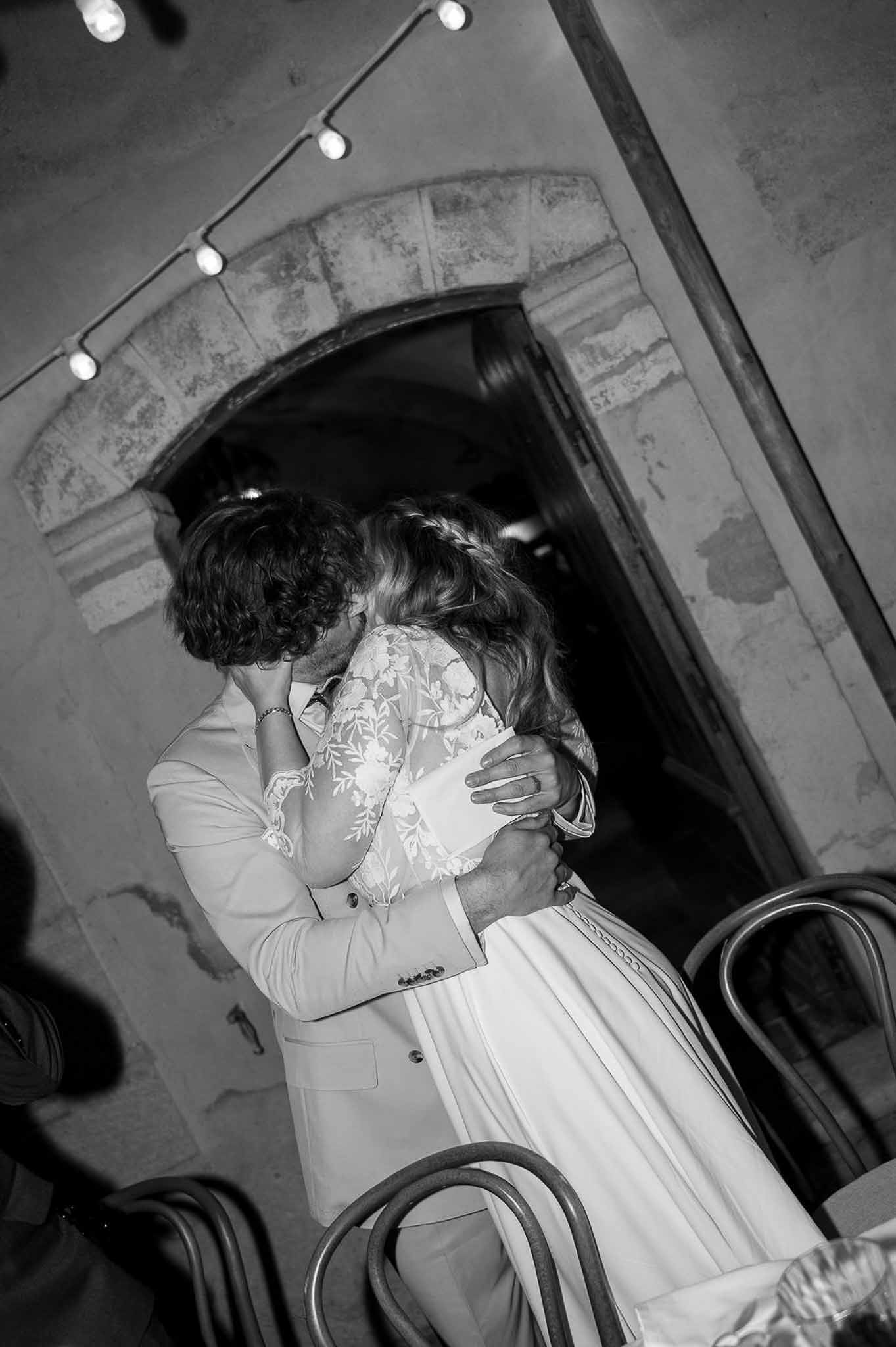 Black and white photo of bride and groom kissing inside a rustic stone venue with arched doorway and bentwood chairs