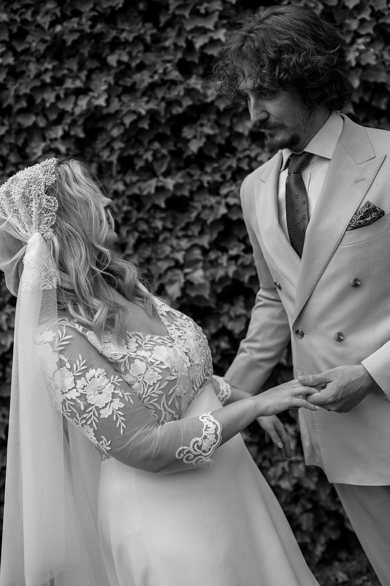 Black and white close-up of bride in embroidered gown and groom in double-breasted suit against ivy wall