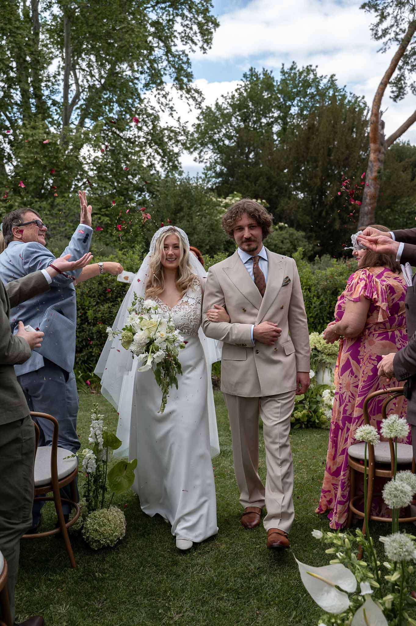 Couple walking down aisle with guests tossing red and pink petals past allium and calla lily markers