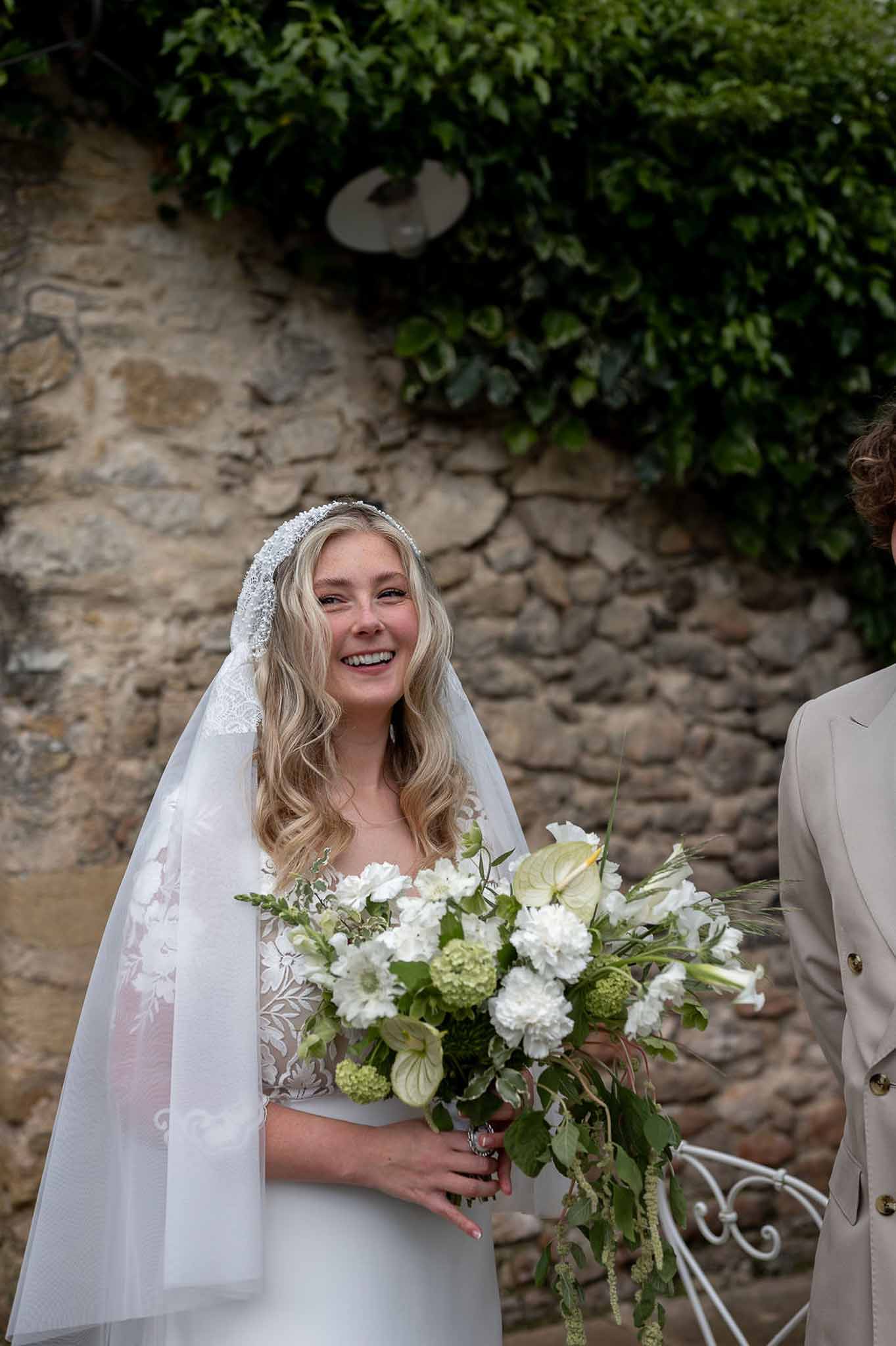 Laughing bride in lace gown and beaded veil holding a bouquet of white dahlias and anthuriums by a stone wall