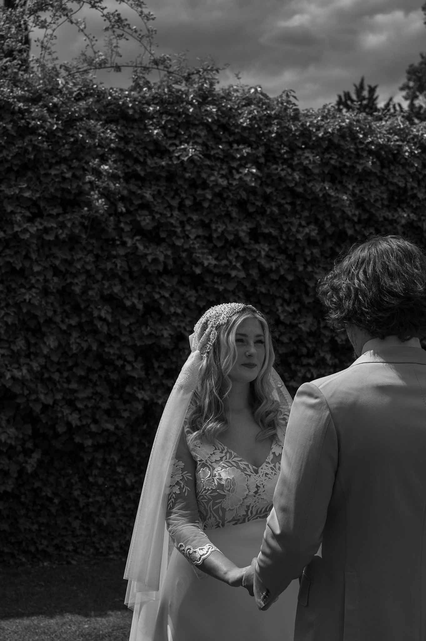 Bride in floral lace gown and embroidered cap veil holding groom's hands before ivy wall in B&W